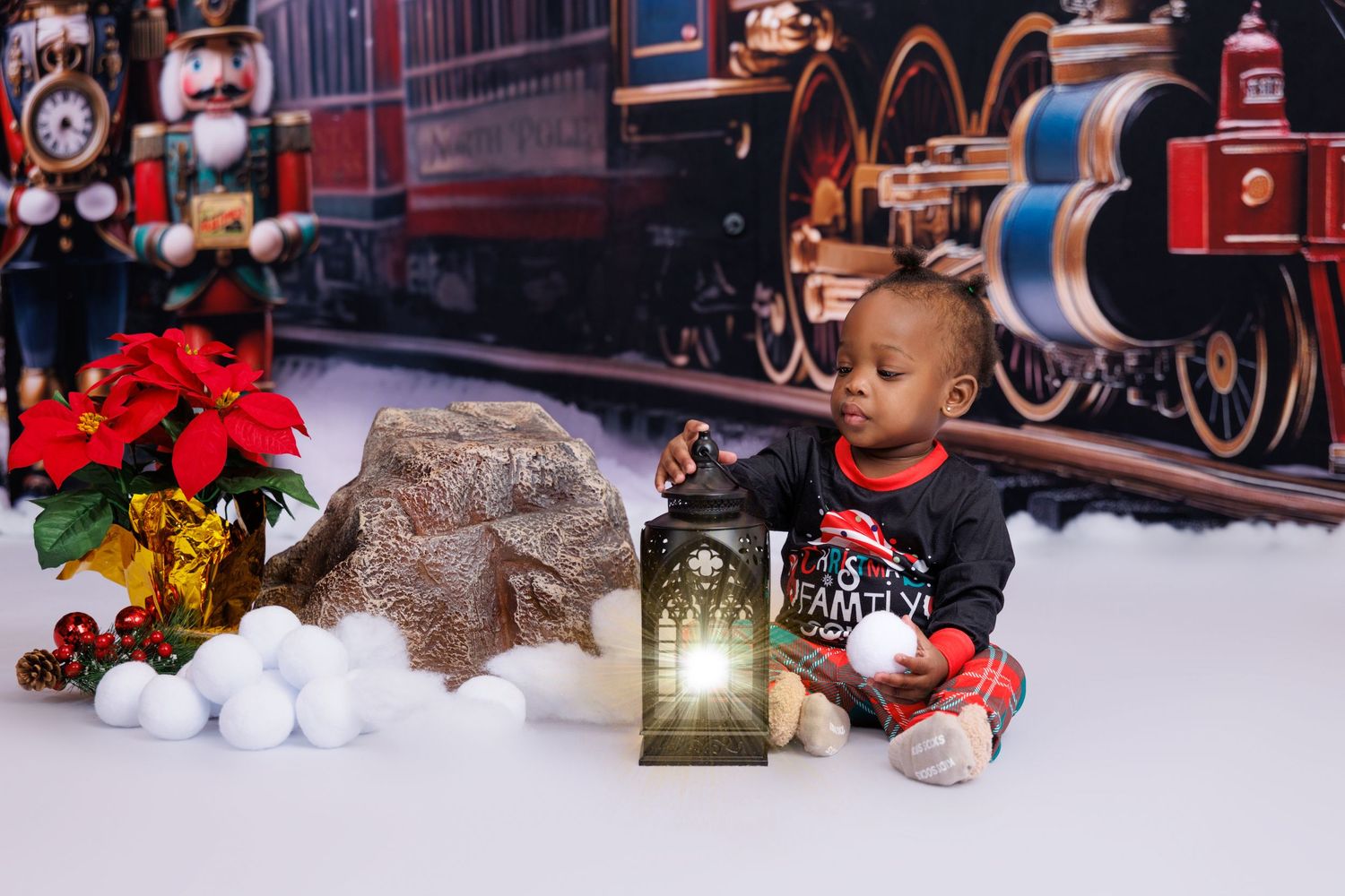 Baby boy in Christmas outfit sits with lantern and snowballs near festive train holiday backdrop.