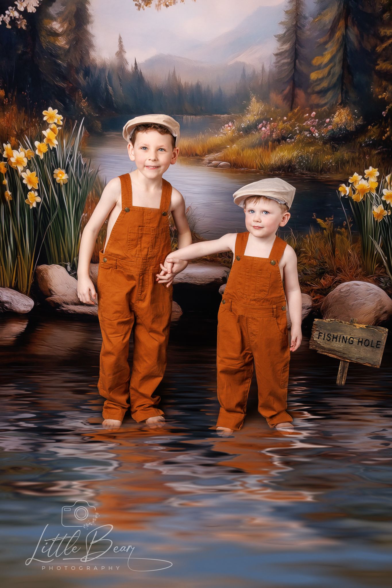 Two young brothers in rust orange overalls and caps standing in a scenic river with daffodils blooming.