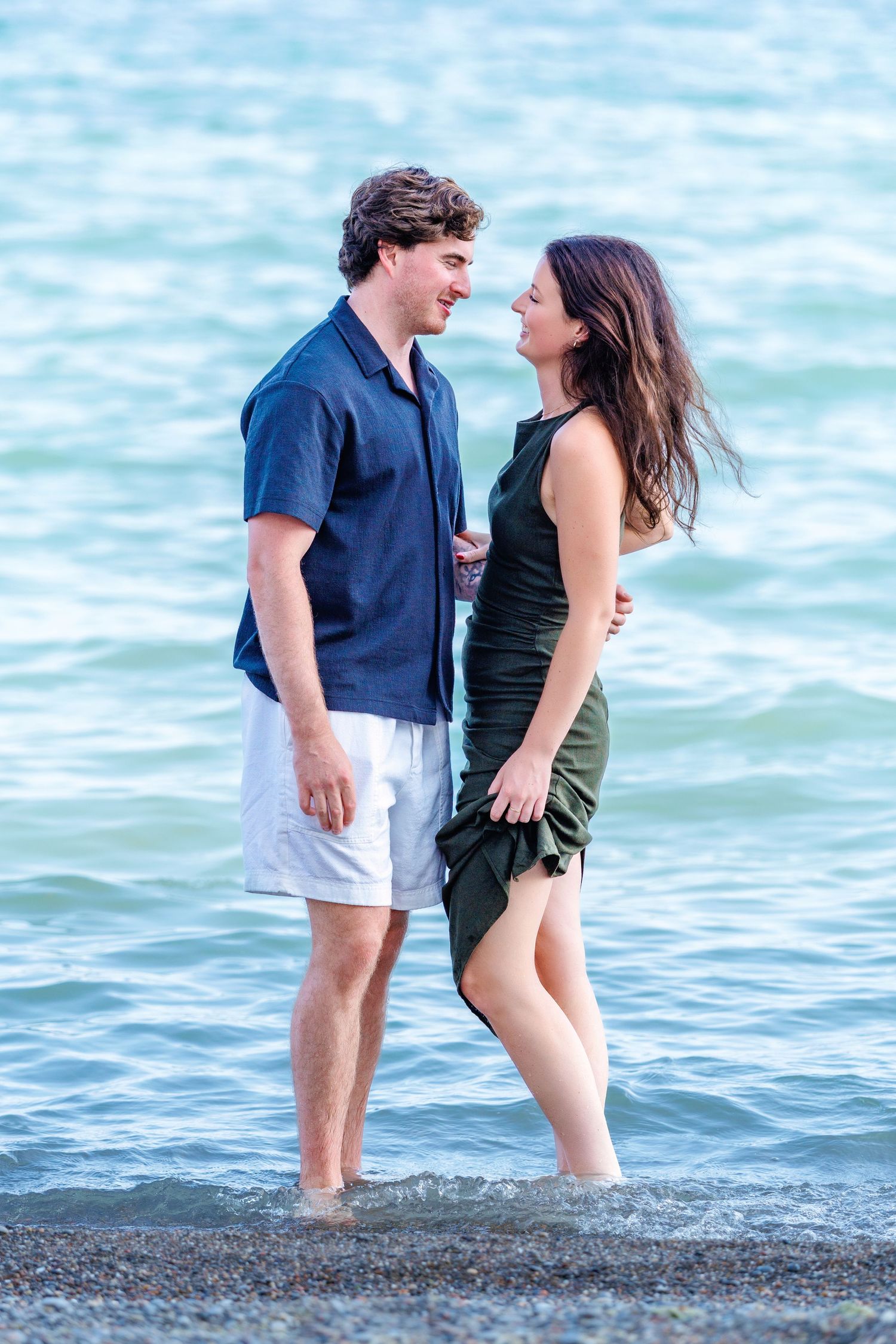 Couple standing in shallow ocean water, smiling at each other, man in navy shirt and woman in green dress.