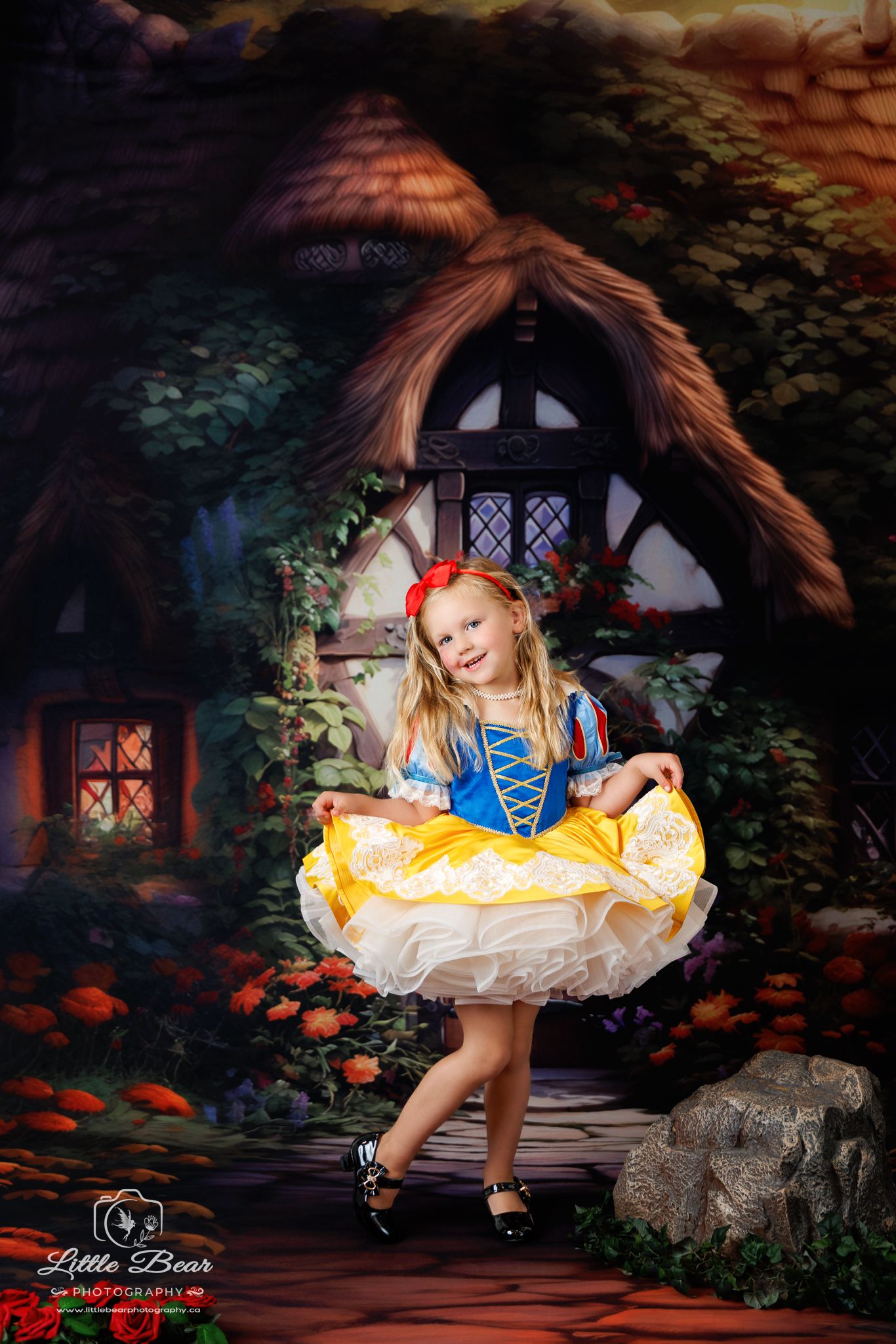 Young girl in Snow White costume poses in front of enchanted cottage backdrop in professional photo.