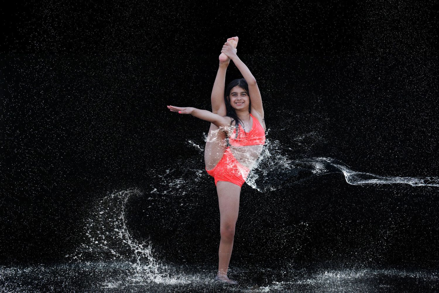 A young woman in a coral bikini performs a graceful dance pose surrounded by dramatic water splashes on a dark background.