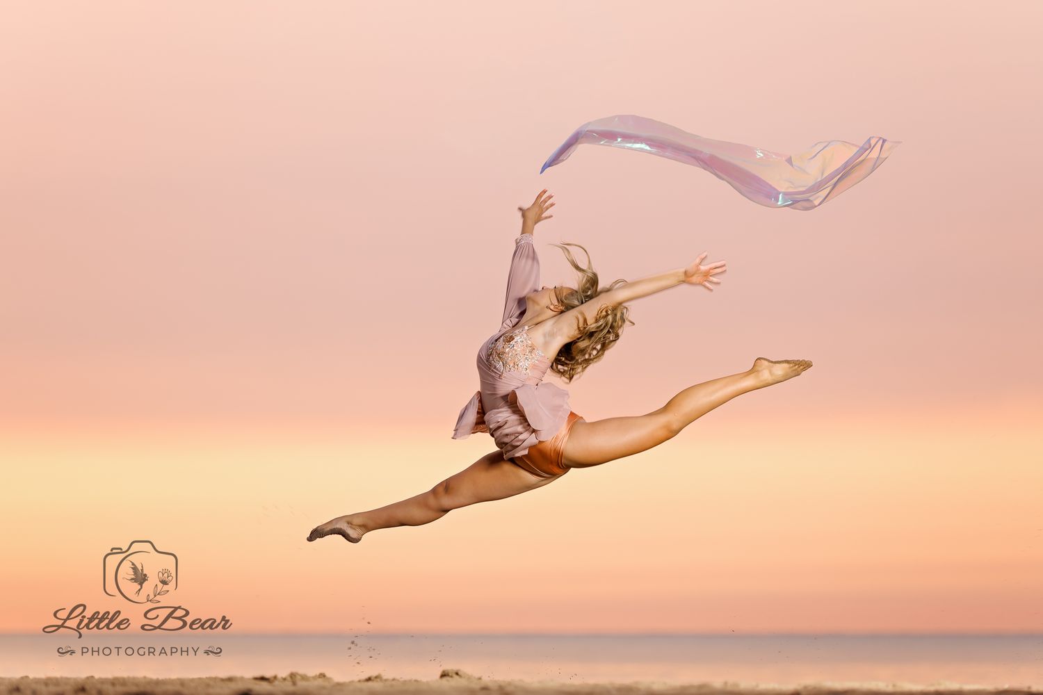 A dancer leaps gracefully mid-air on a beach at sunset, extending a flowing fabric ribbon against a pink sky.