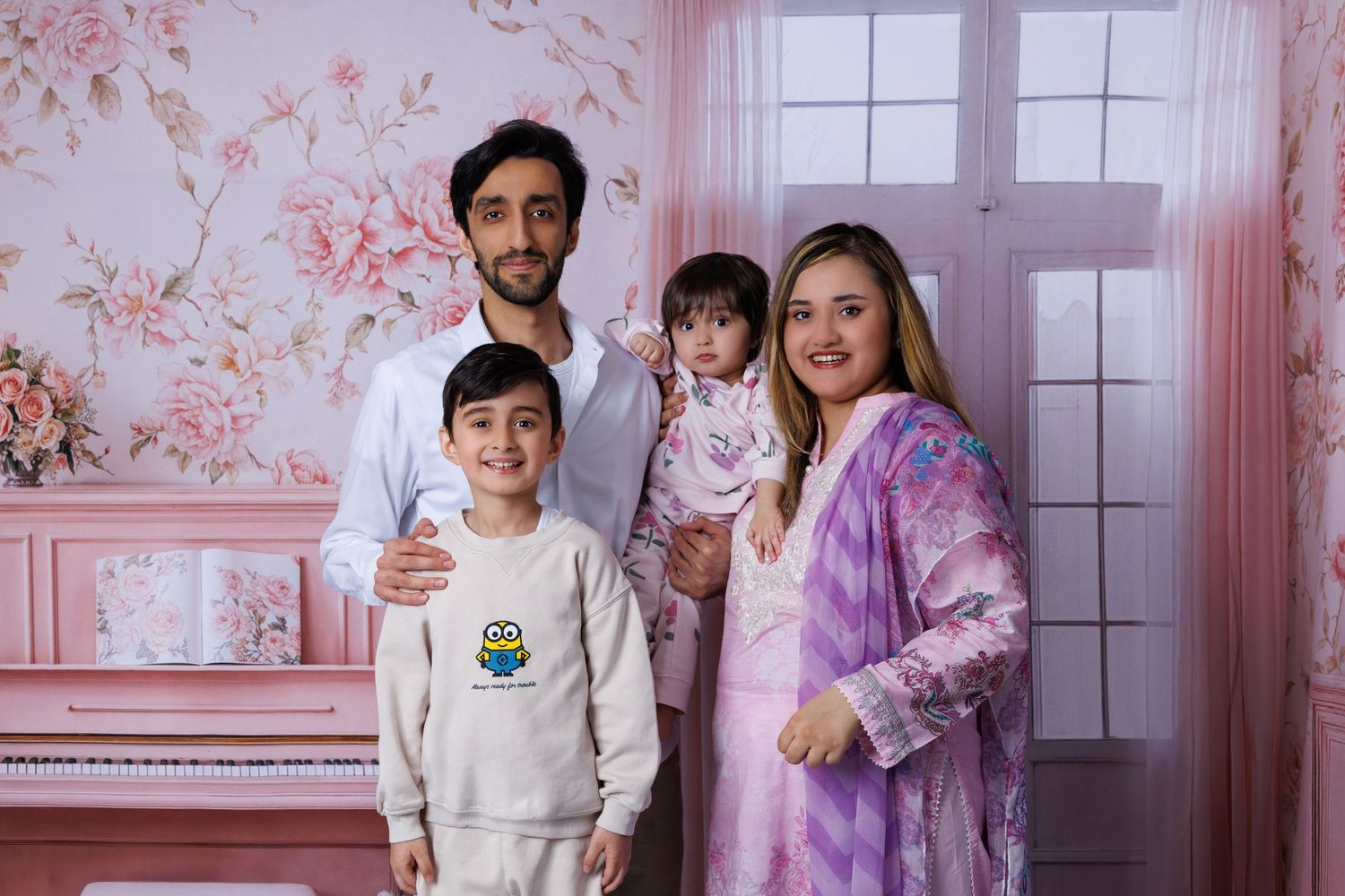 Smiling family of four stands together in pink floral studio with pink piano in background.