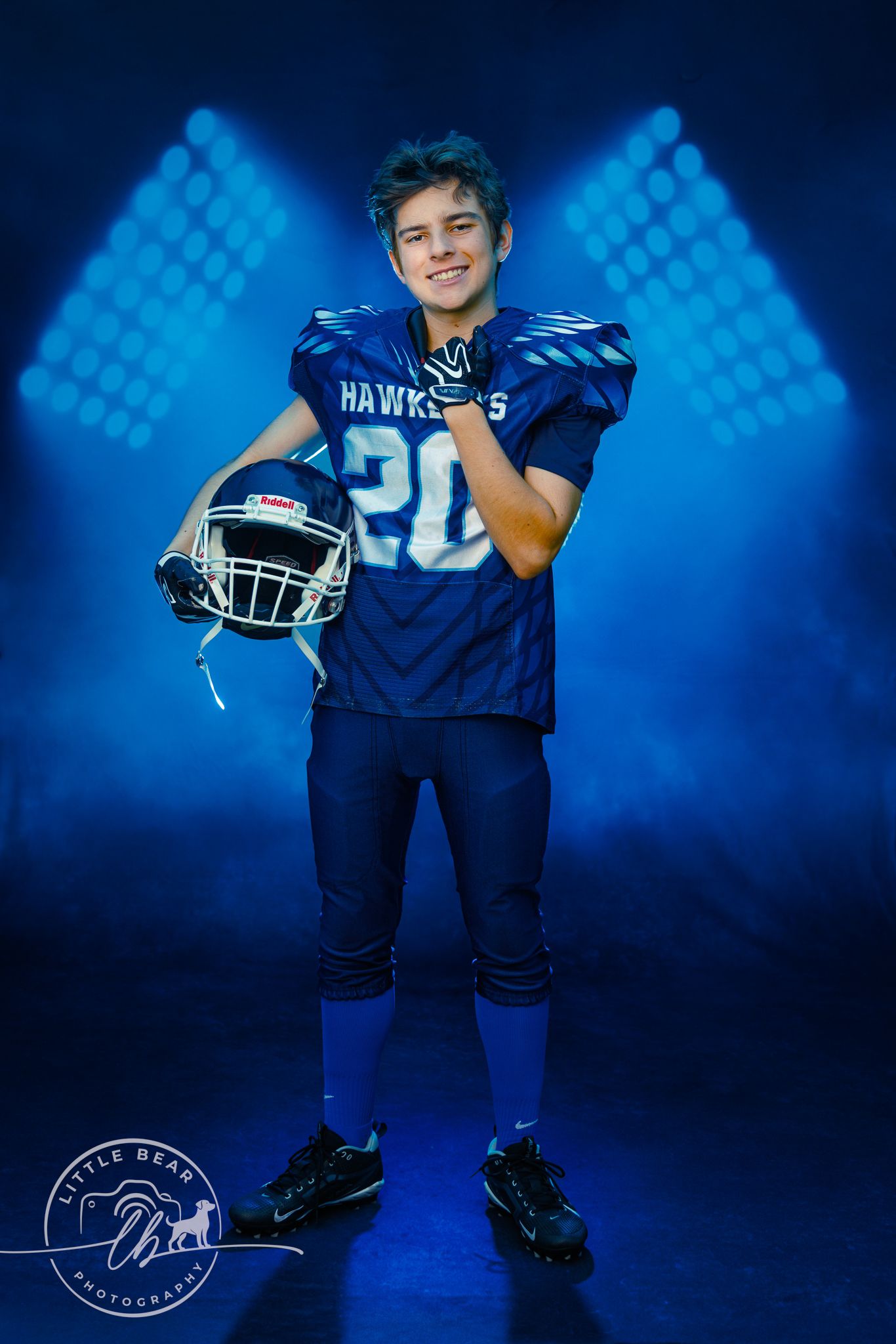 Smiling young Hawkeyes football player number 20 holding helmet under blue stadium lights with smoky background.