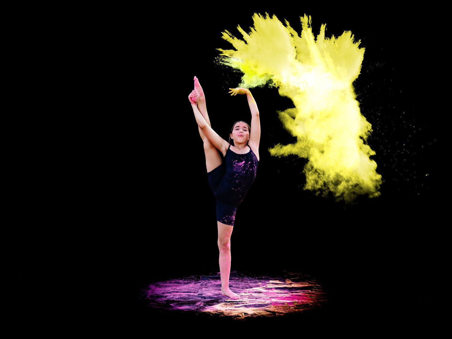 Gymnast executes high leg extension pose with vivid yellow powder cloud on black background.