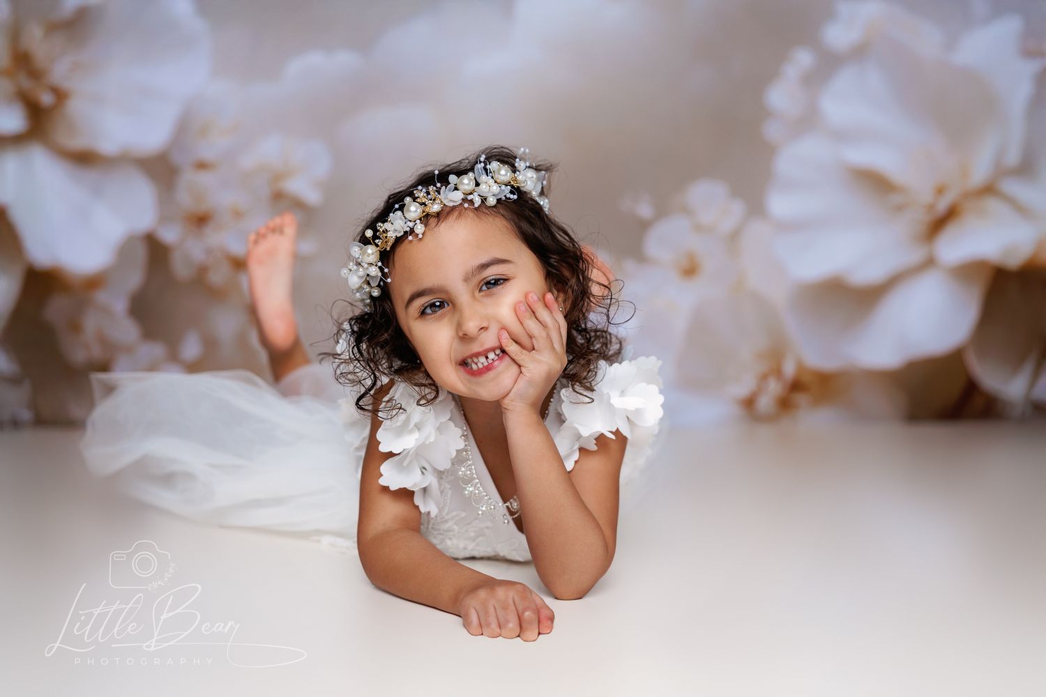 Young girl in white dress and floral crown poses playfully against a white floral backdrop.