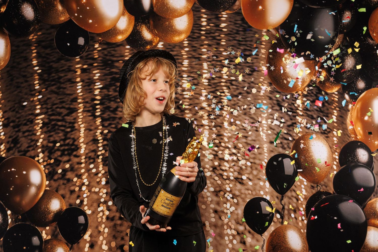 A child in black holds a champagne bottle amid gold and black balloons and falling confetti at a celebration.
