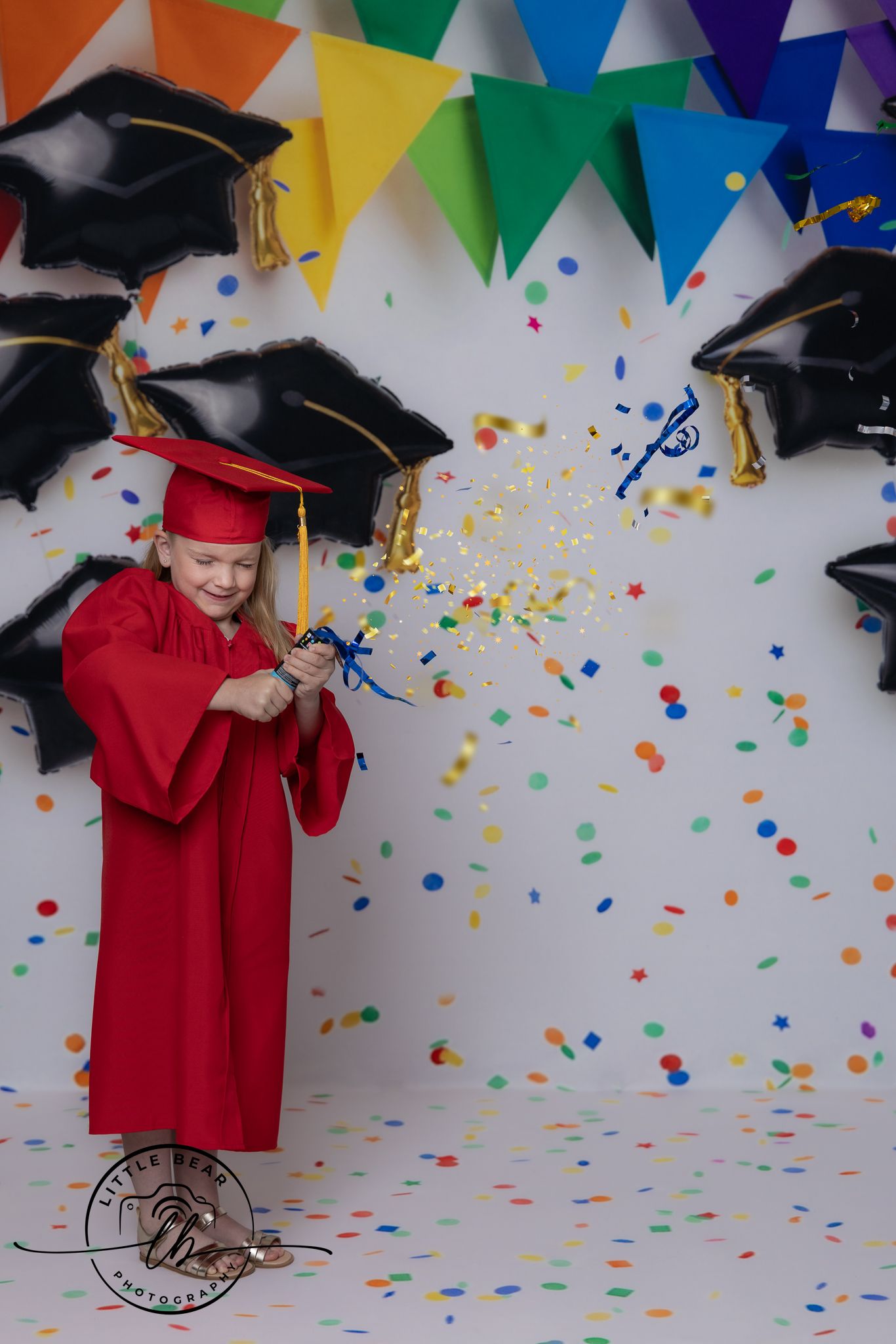 Young girl in red graduation cap and gown popping a confetti cannon, surrounded by colorful balloons and bunting.