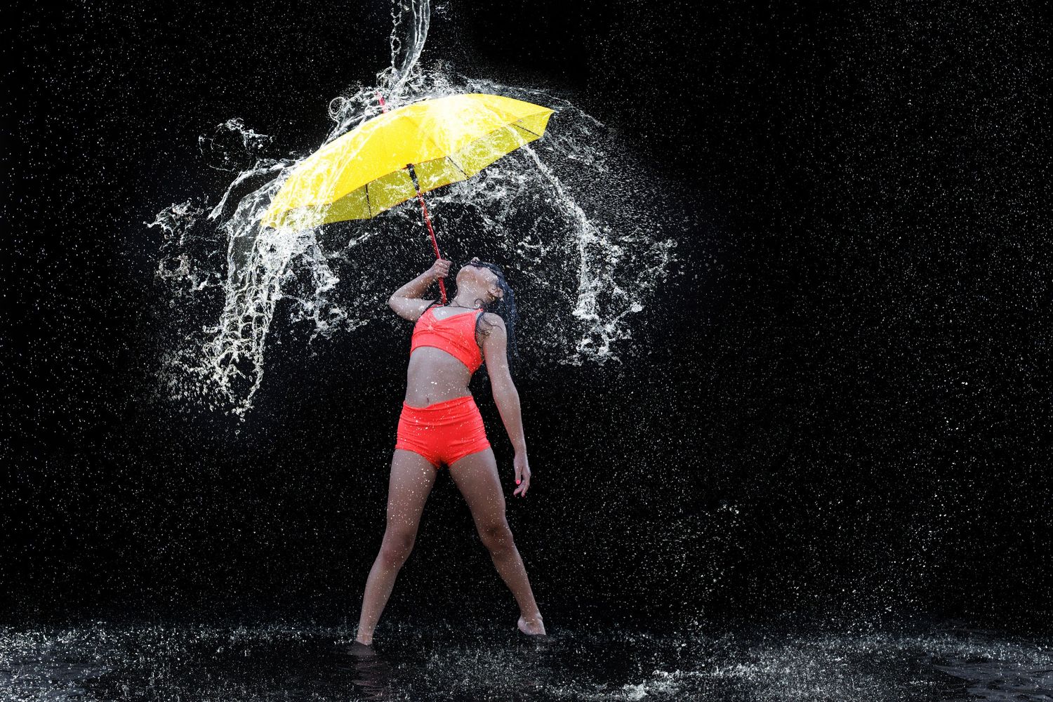 Woman in orange bikini holding yellow umbrella amid dramatic water splashes against black background.