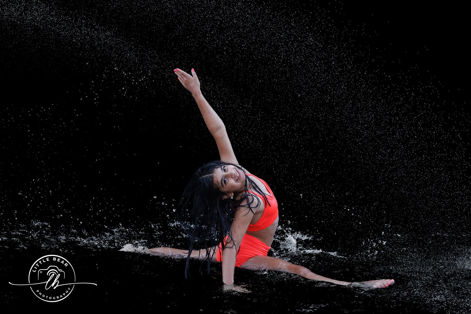 Dancer in red leotard performs dramatic split leap surrounded by water splashes against black background.