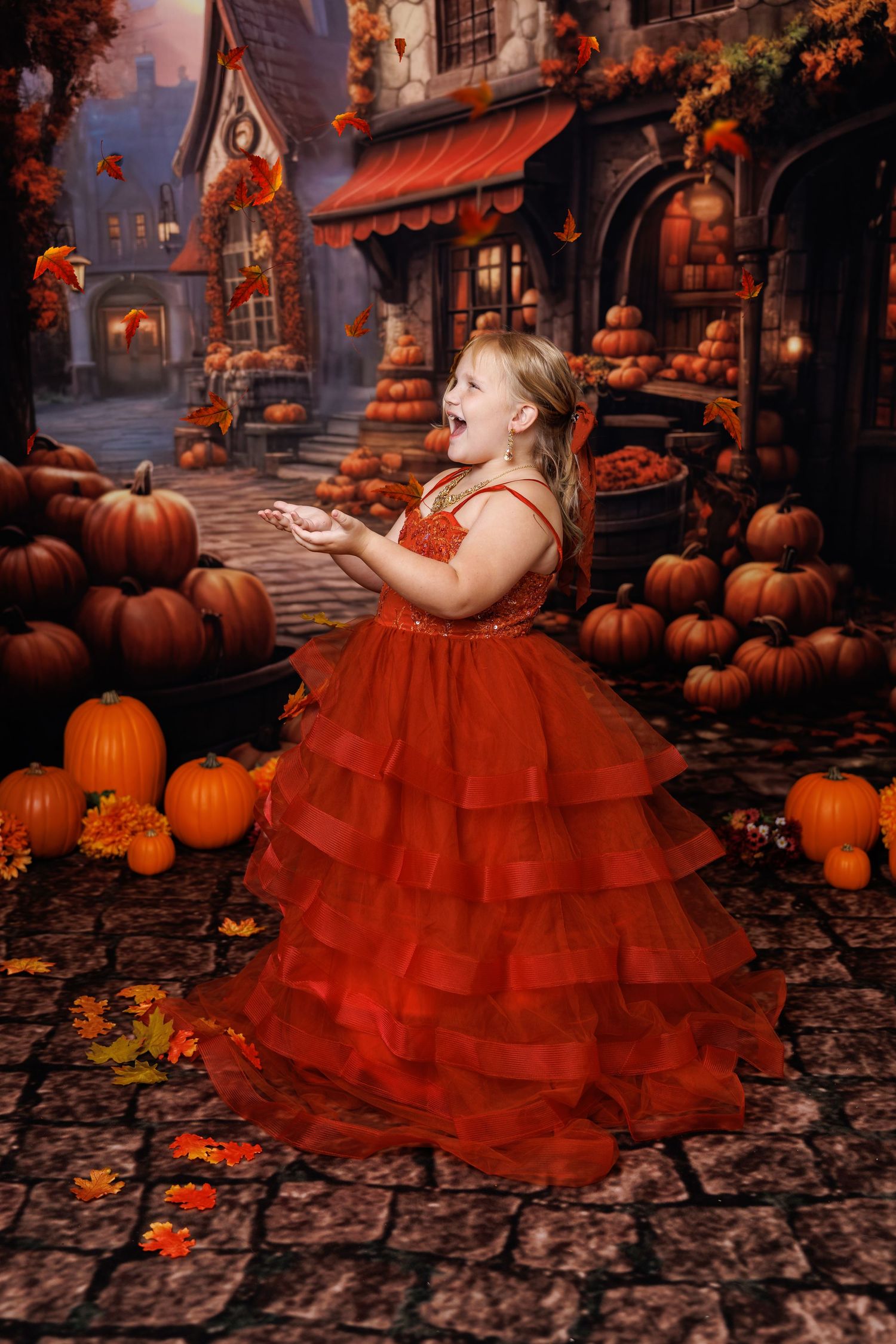 A joyful girl in a red ruffled gown poses among pumpkins in a festive autumn Halloween village setting.