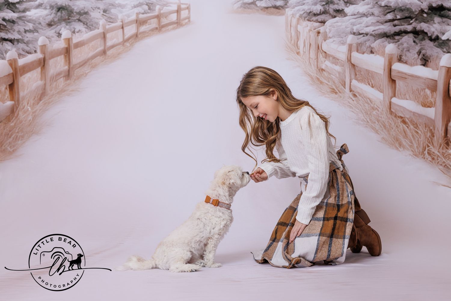 A young girl in a plaid skirt feeds a white fluffy dog in a snowy winter scene with white fences and trees.