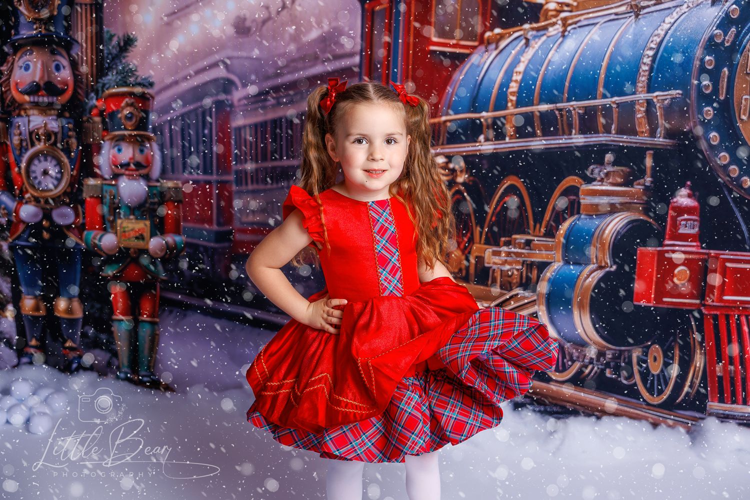 Young girl in red plaid dress posing before Christmas train and nutcracker photography backdrop with snow.
