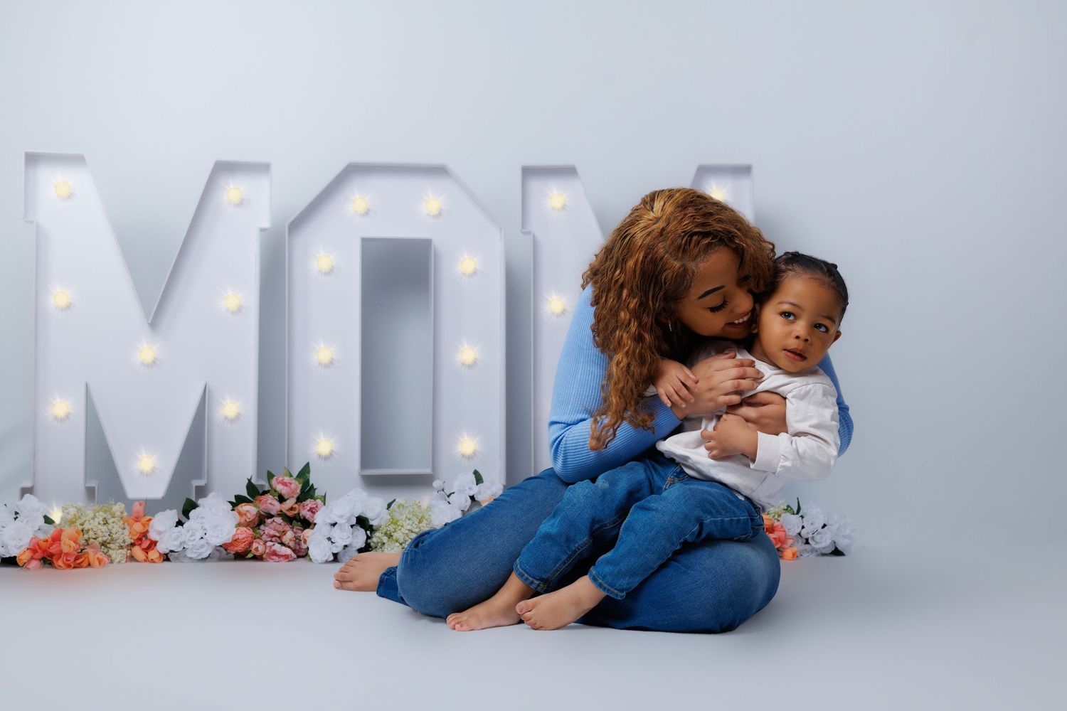 A mother tenderly embraces her toddler in front of illuminated MOM letters with floral decorations.