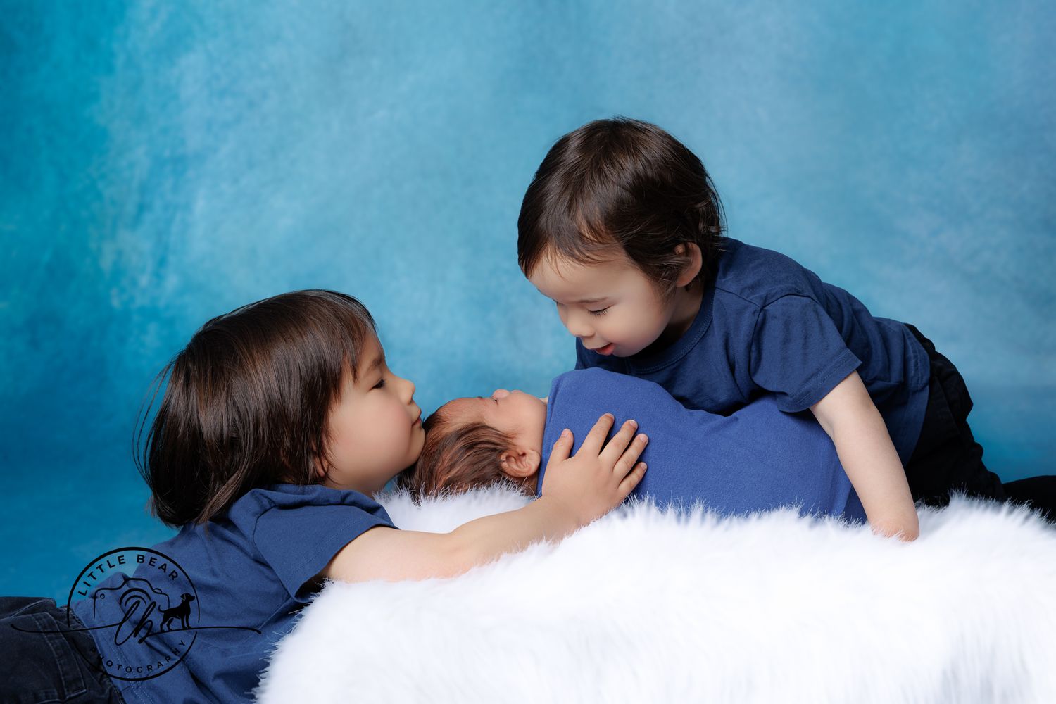 Two young siblings in matching blue outfits tenderly interact with a newborn baby on a white fluffy surface.