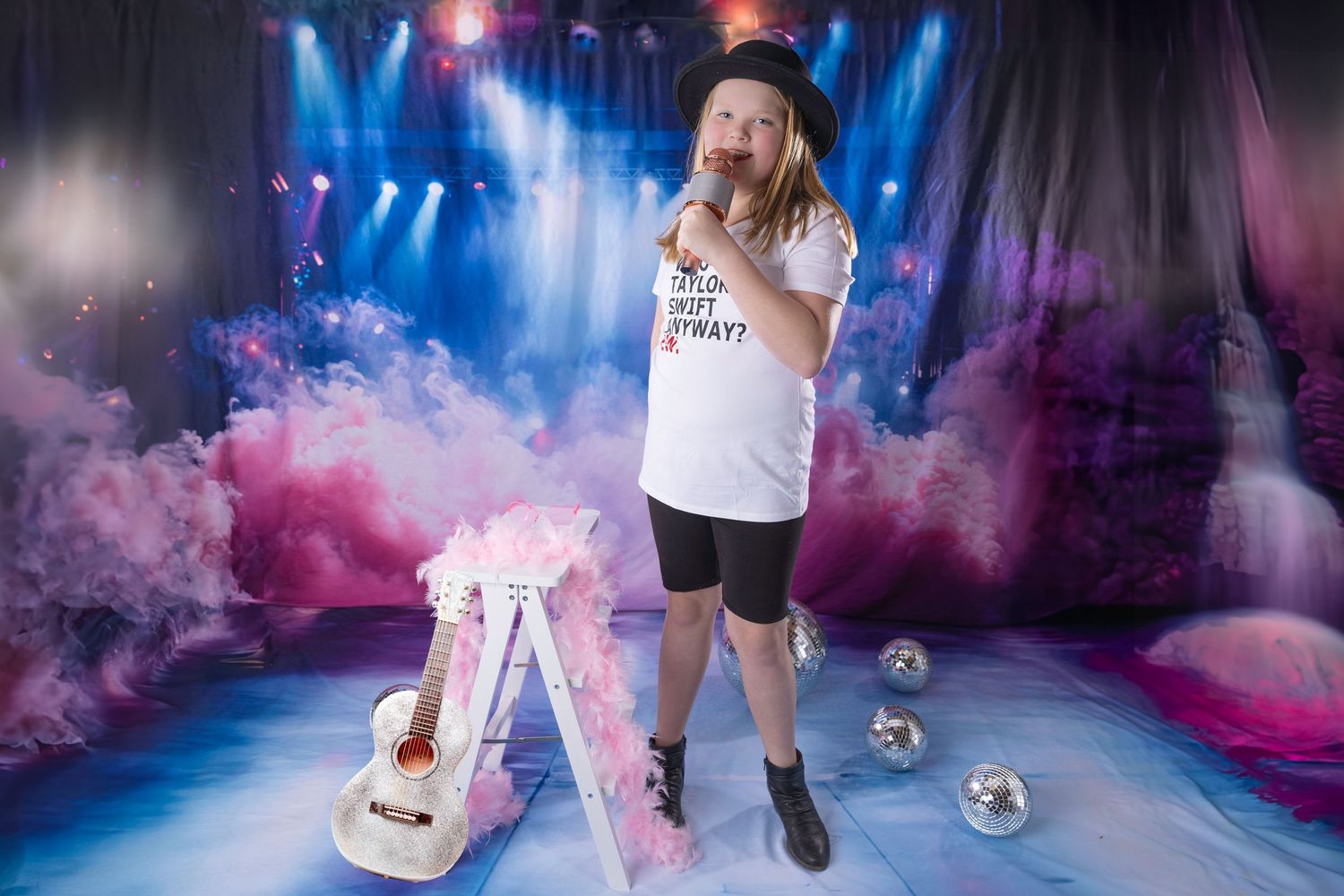 Young performer in black hat and Taylor Swift tee holds microphone on colorful smoke-filled stage with guitar and disco balls.
