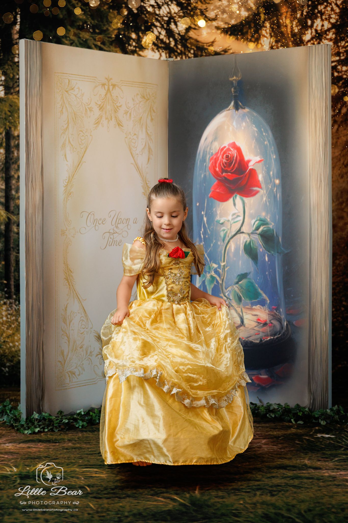 Young girl in Belle yellow gown poses before Beauty and the Beast storybook backdrop with enchanted rose.