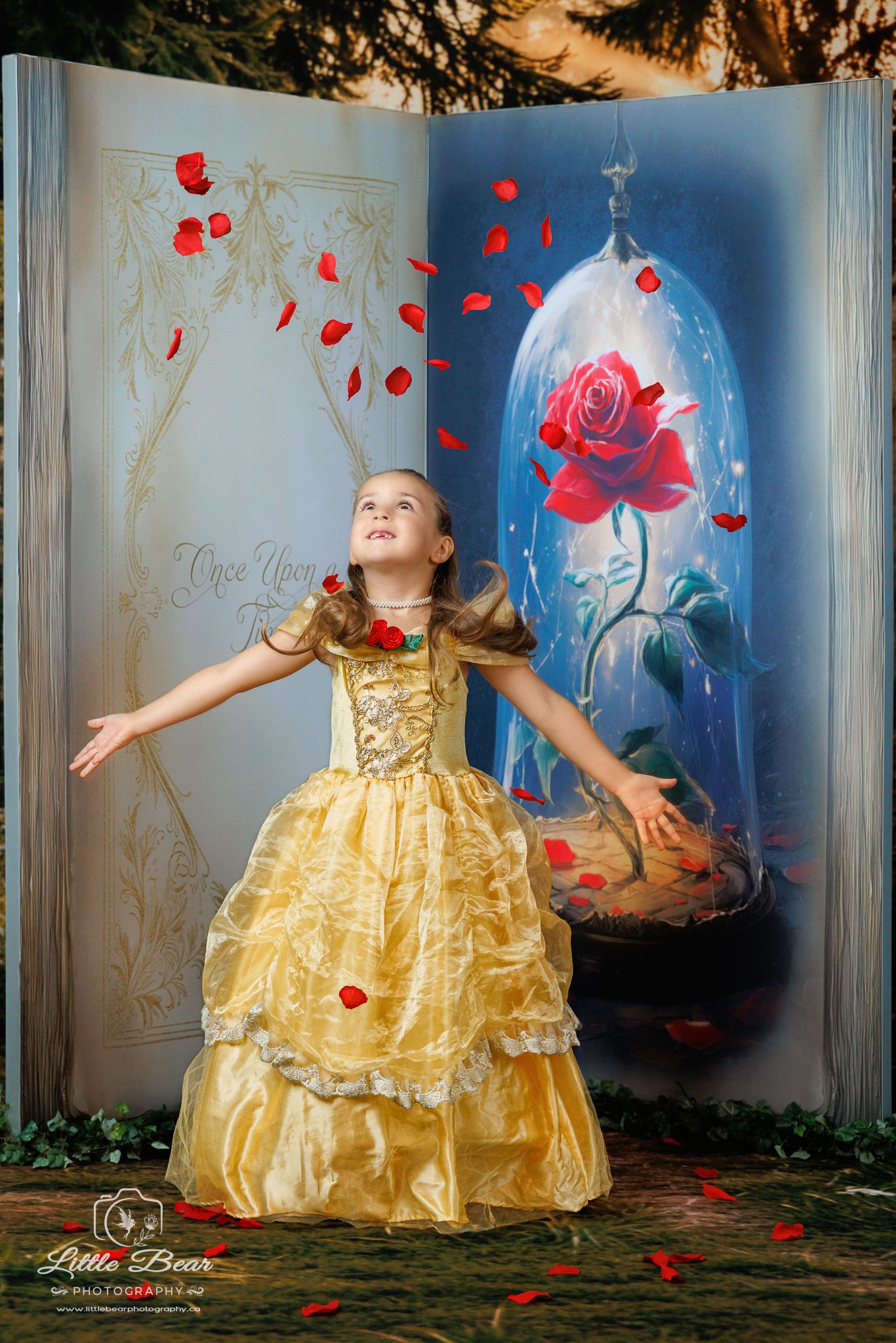 Girl in gold Belle gown poses joyfully before enchanted rose backdrop with red petals falling around her.
