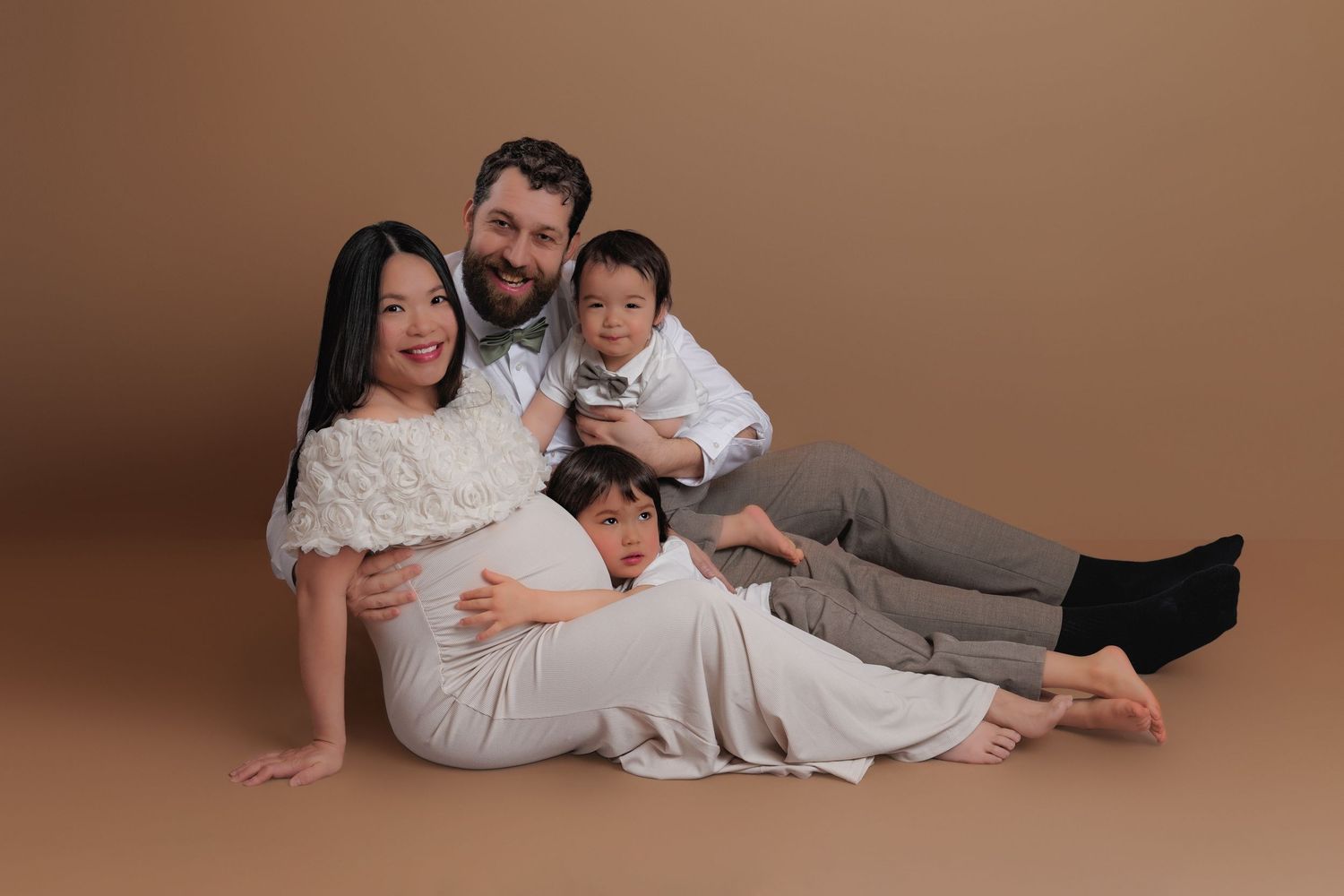 Joyful family of four with pregnant mother sitting casually together against warm brown studio backdrop.