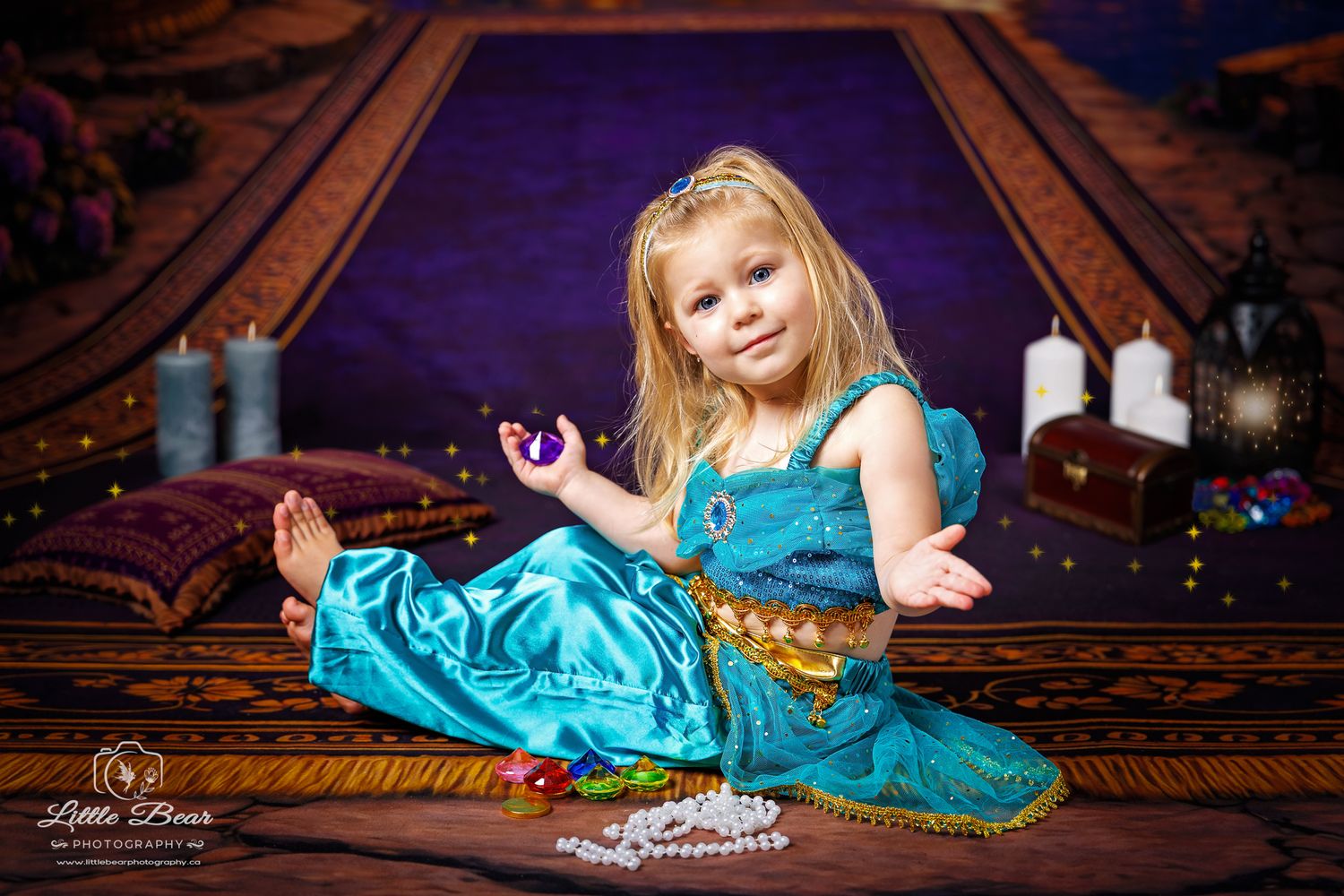 Young girl in teal princess costume sits on decorative rug with candles and treasure chest in magical photo shoot.