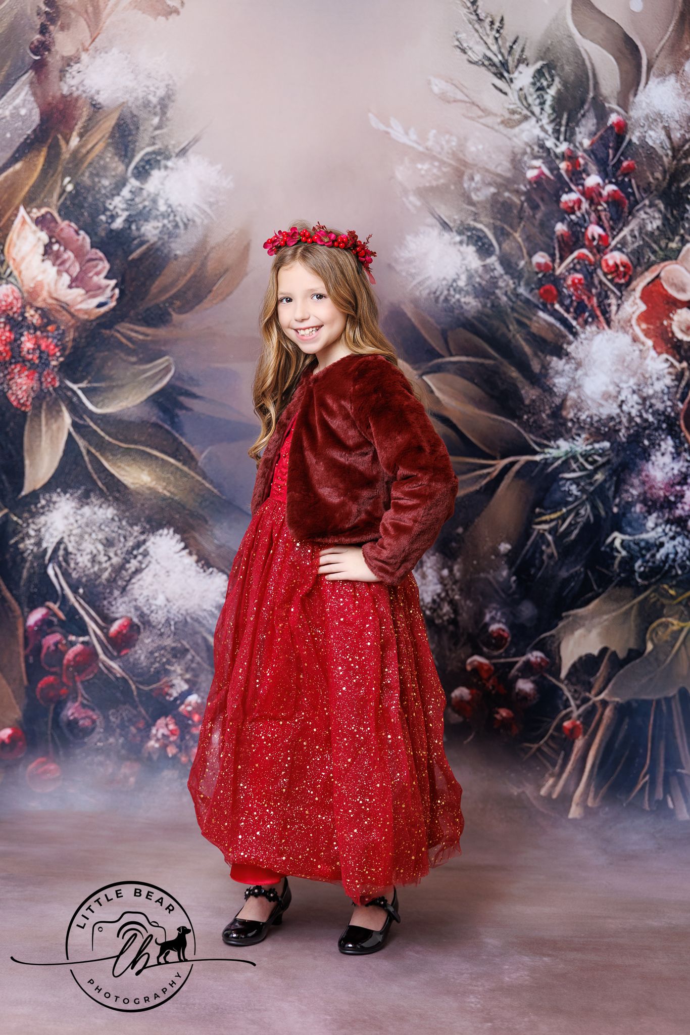 A young girl in a red sparkly dress and floral crown poses before a festive winter floral backdrop.