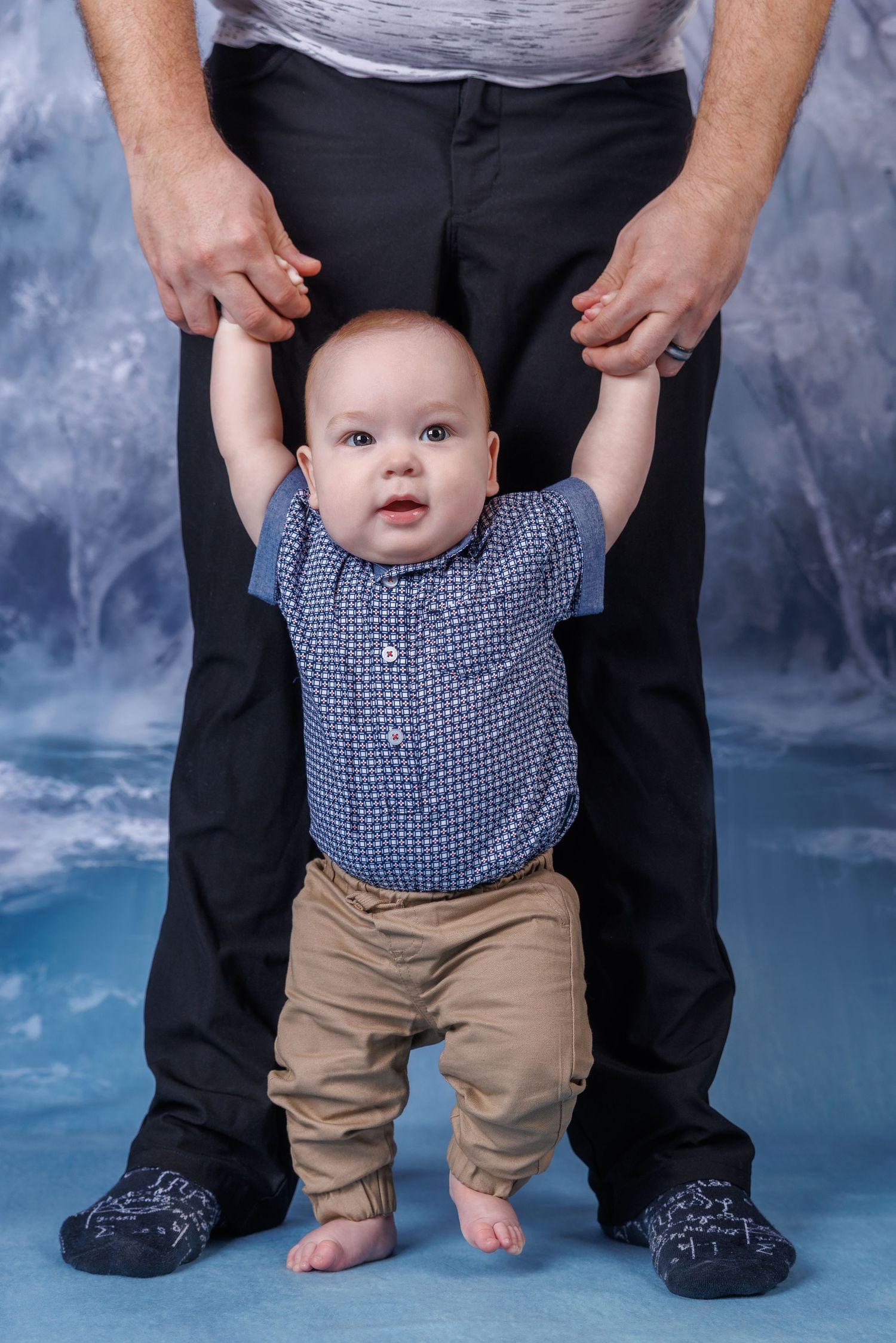 Curious baby boy in blue shirt stands supported by adult hands against a blue studio backdrop.