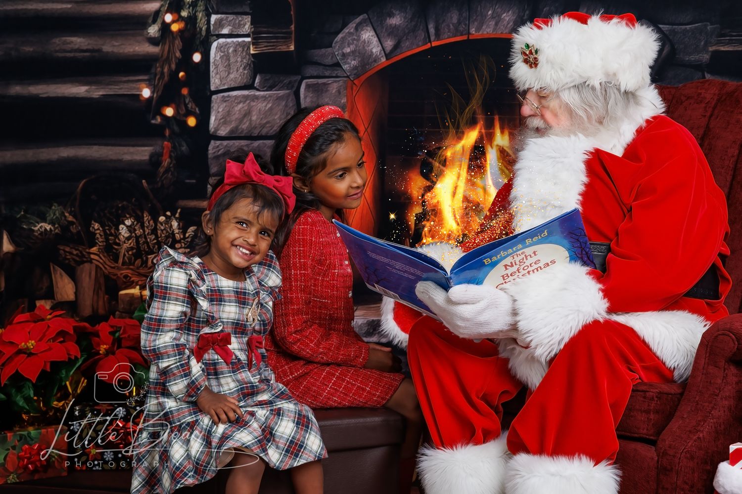 Two children sit with Santa Claus reading a Christmas book by a cozy fireplace with holiday decorations.