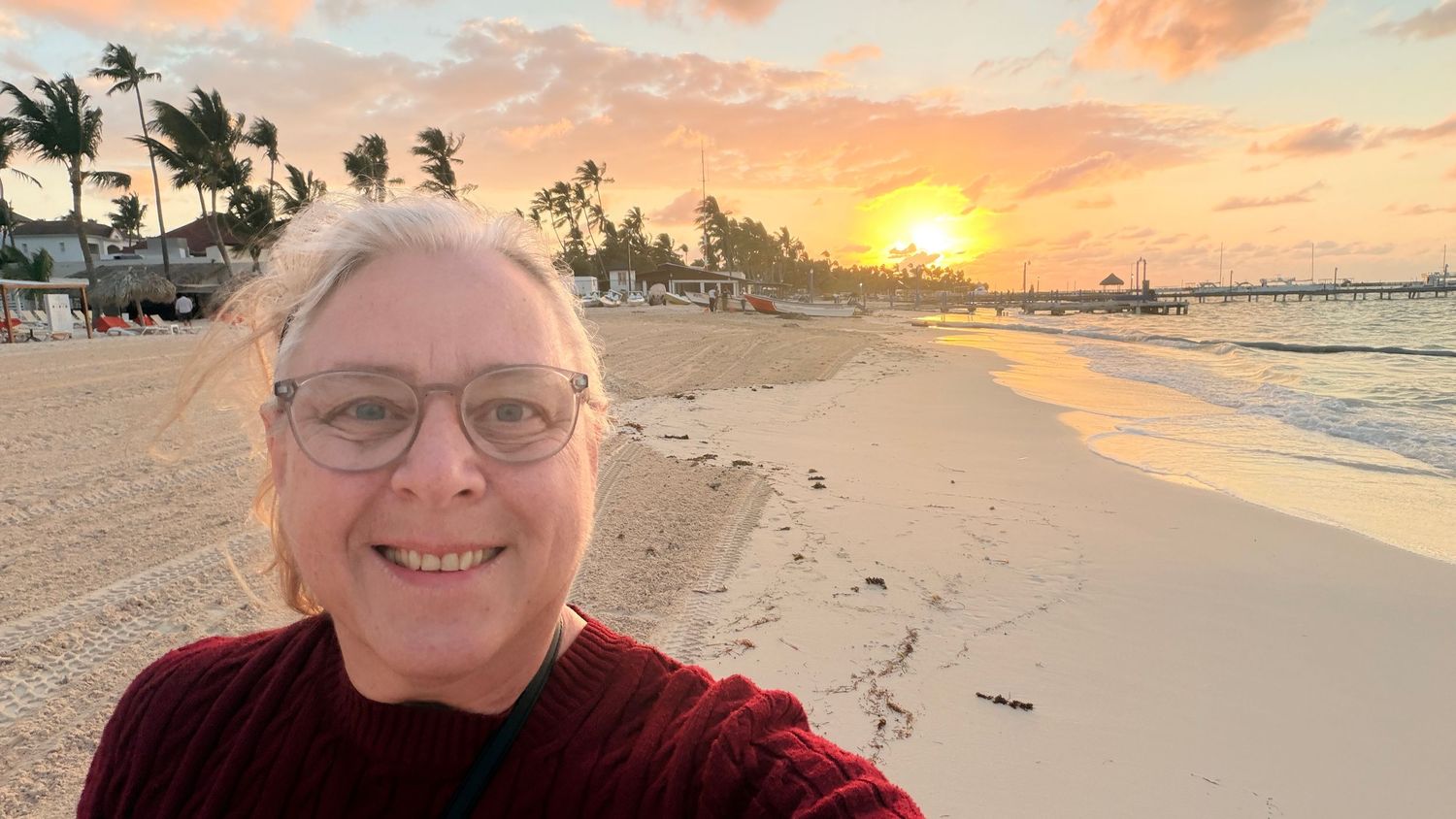 Smiling woman with glasses takes a beach selfie at sunset with palm trees and golden sky in a tropical destination.