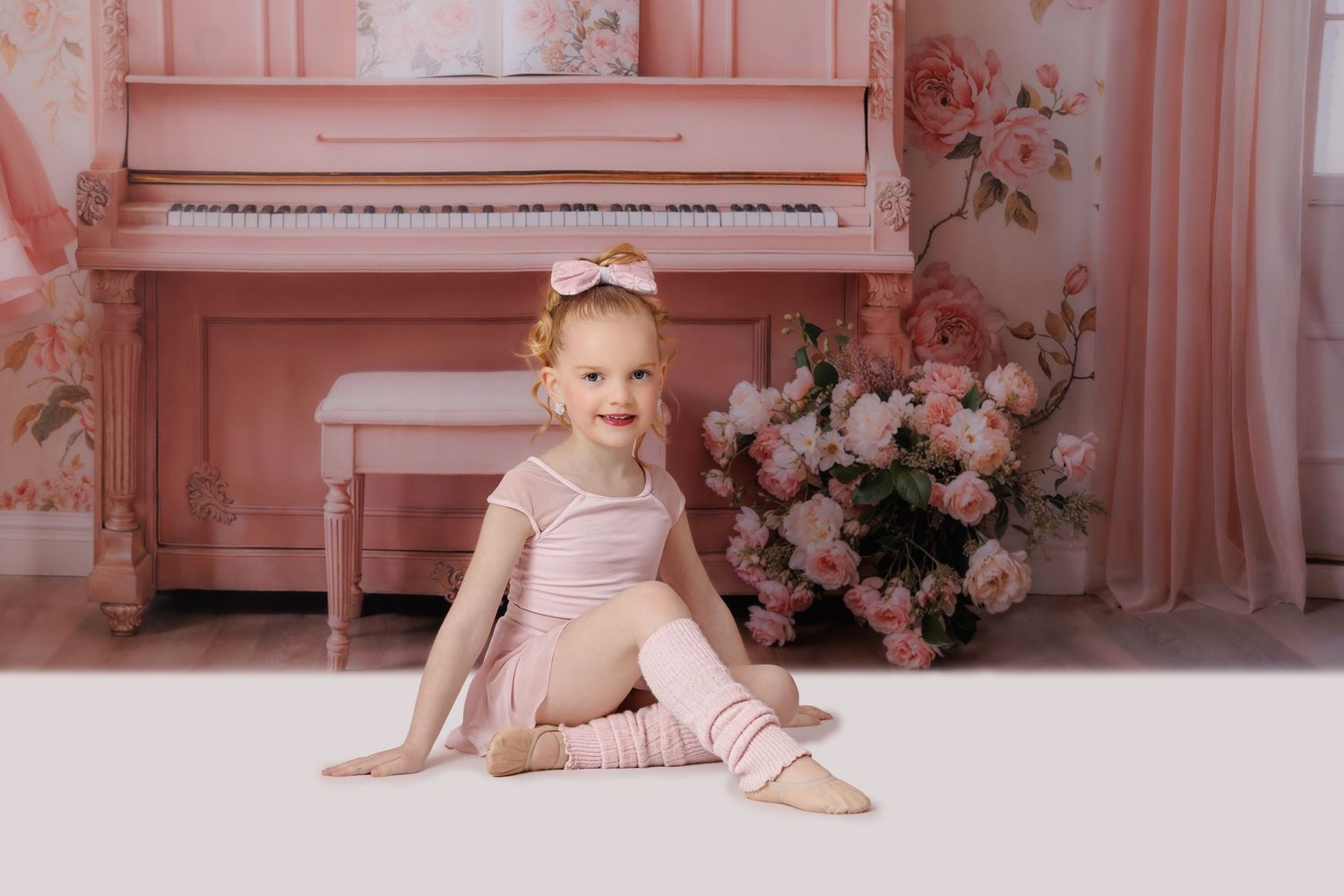 Toddler girl in pink ballet outfit sitting in front of a pink piano with floral wallpaper backdrop.