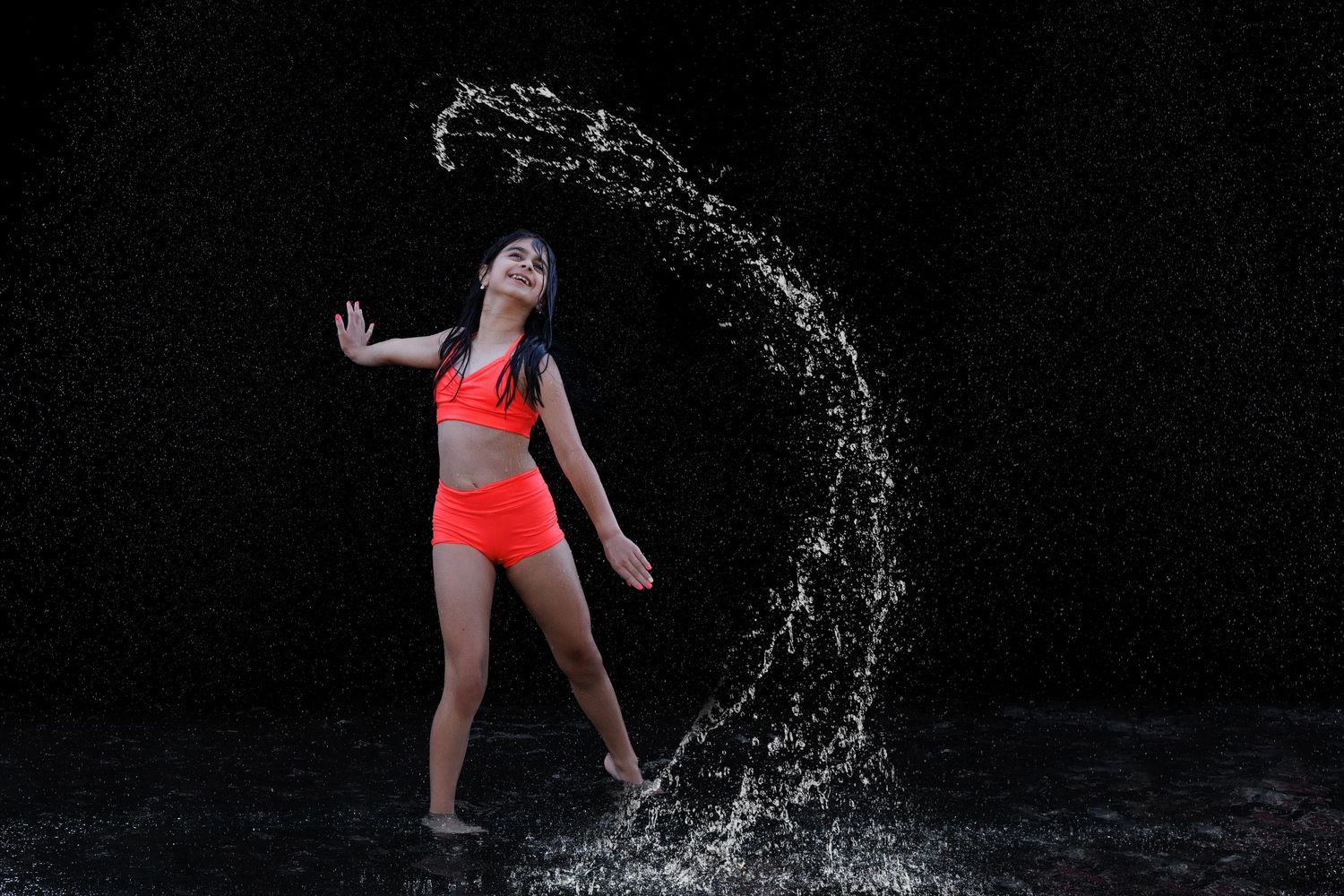 Girl in red bikini poses dramatically as a curved water splash arcs around her against a dark background.