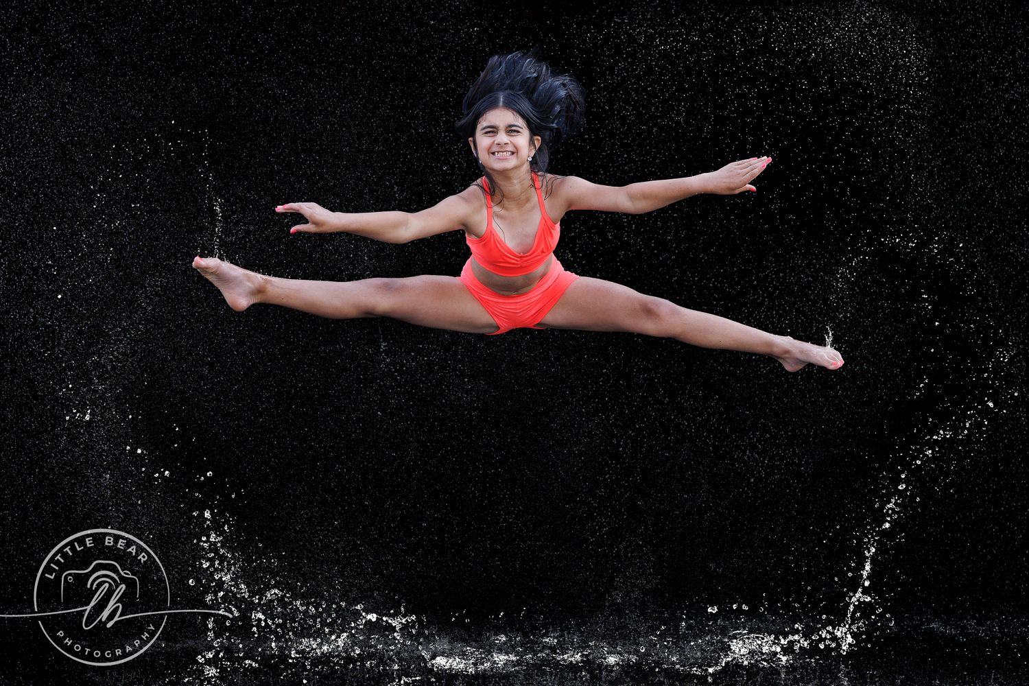 Dancer leaping mid-air in red swimsuit, performing a perfect split against a black background with water splashes.