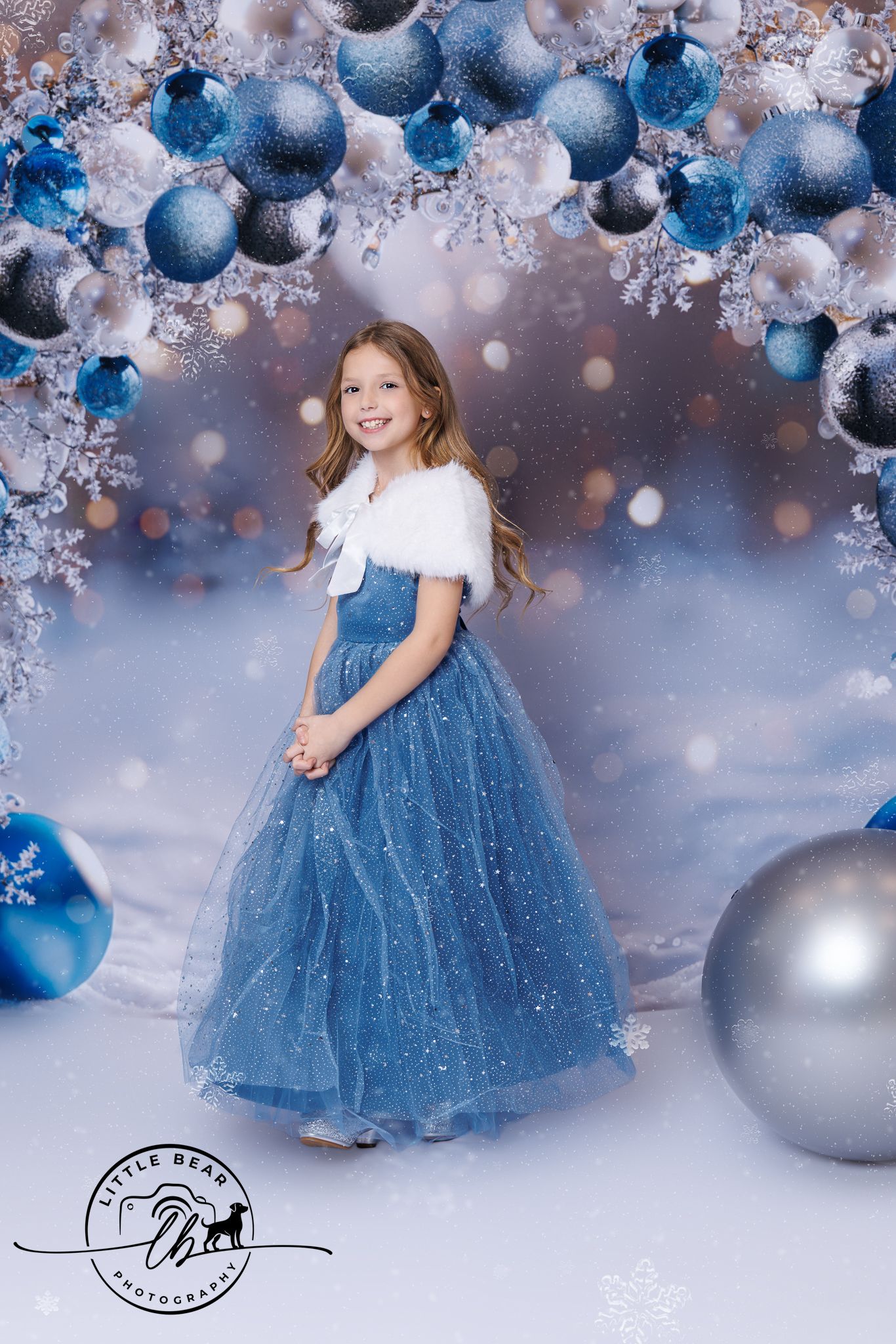 Young girl in blue tulle gown and white fur shrug poses among blue Christmas ornaments in a winter wonderland setting.
