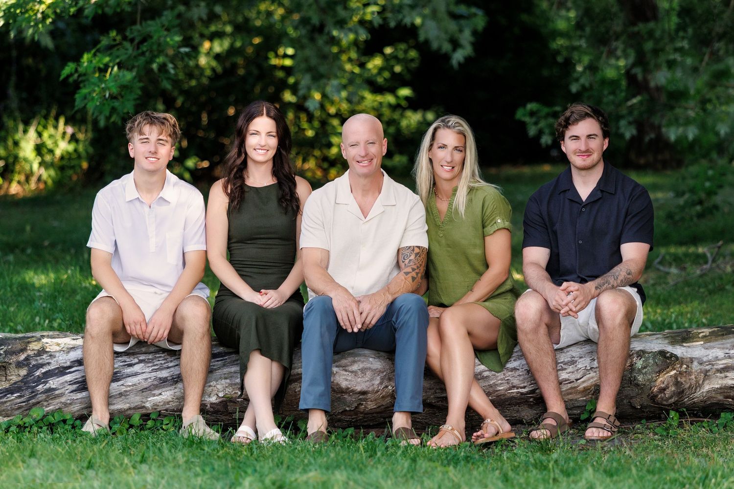 A happy family of five poses together on a fallen log in a lush green park setting.