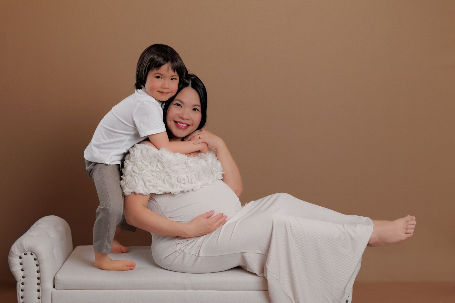 Pregnant mother in white gown reclining on bench while young son lovingly embraces her from behind.