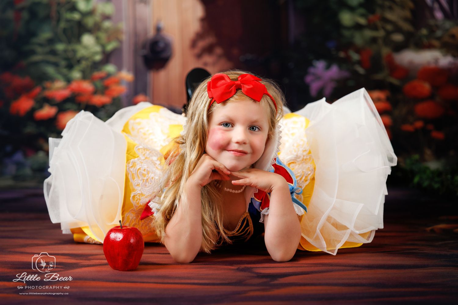 Young girl in Snow White costume with red bow poses with apple on wooden floor before colorful backdrop.