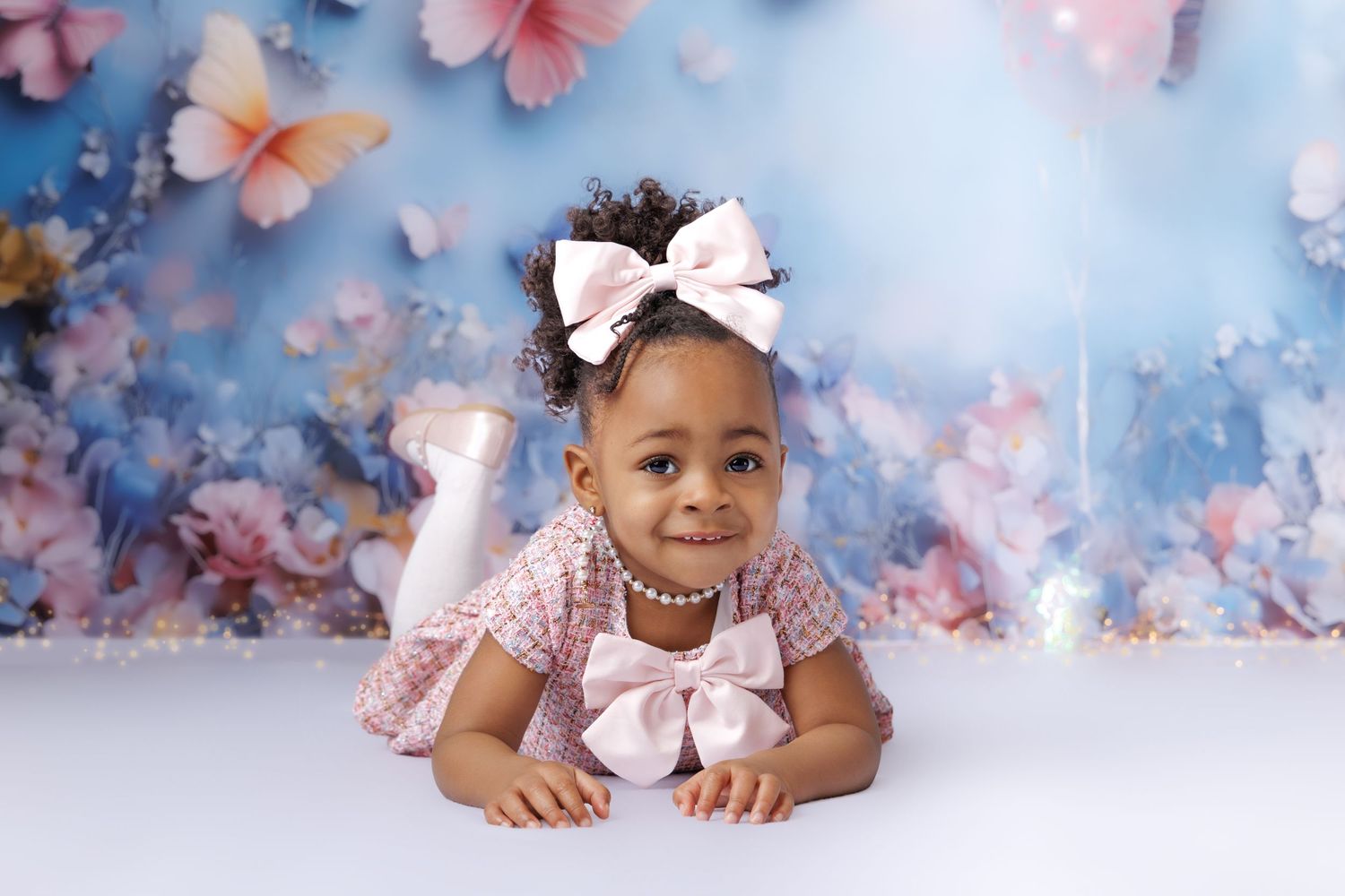 Adorable baby girl in pink bow headband and pearl necklace poses on floral butterfly backdrop.