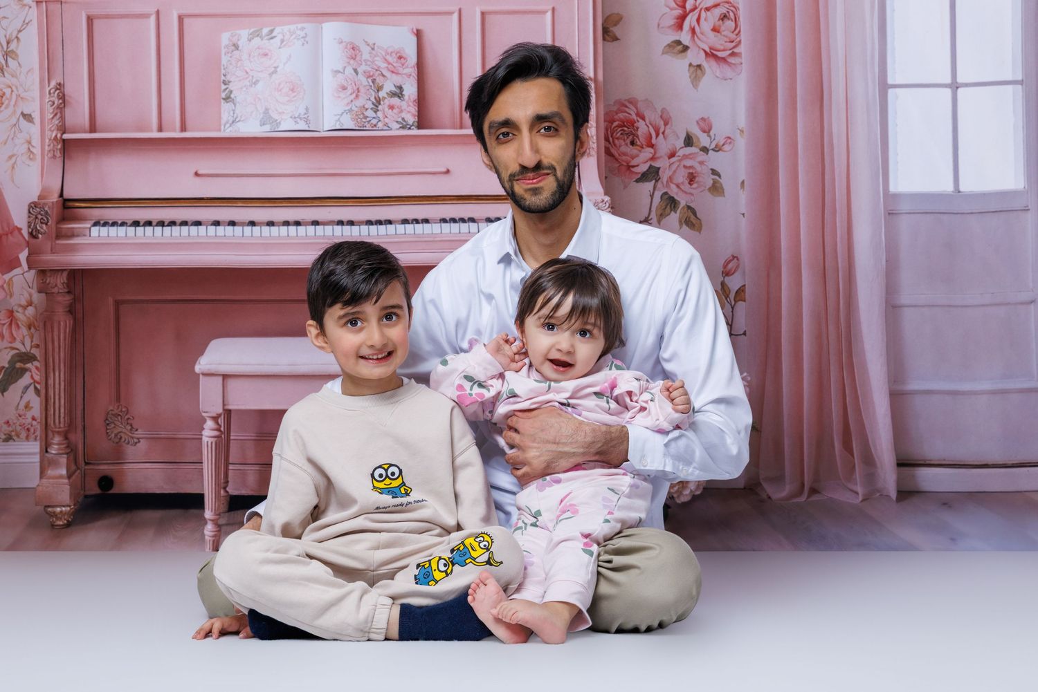 A man sits cross-legged with a toddler boy and baby girl in a pink floral studio setting.