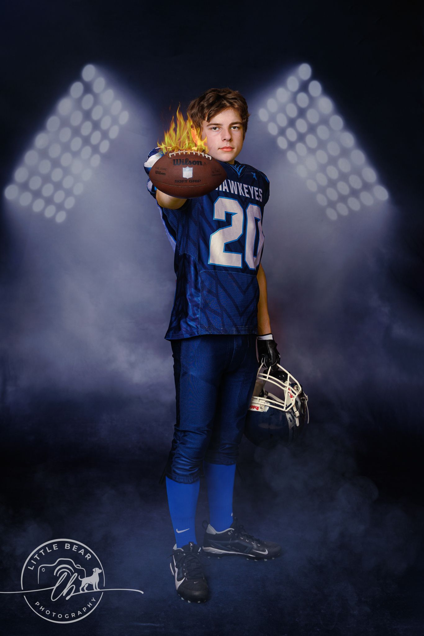 Young Hawkeyes football player #20 holding flaming football and helmet under dramatic stadium spotlights.