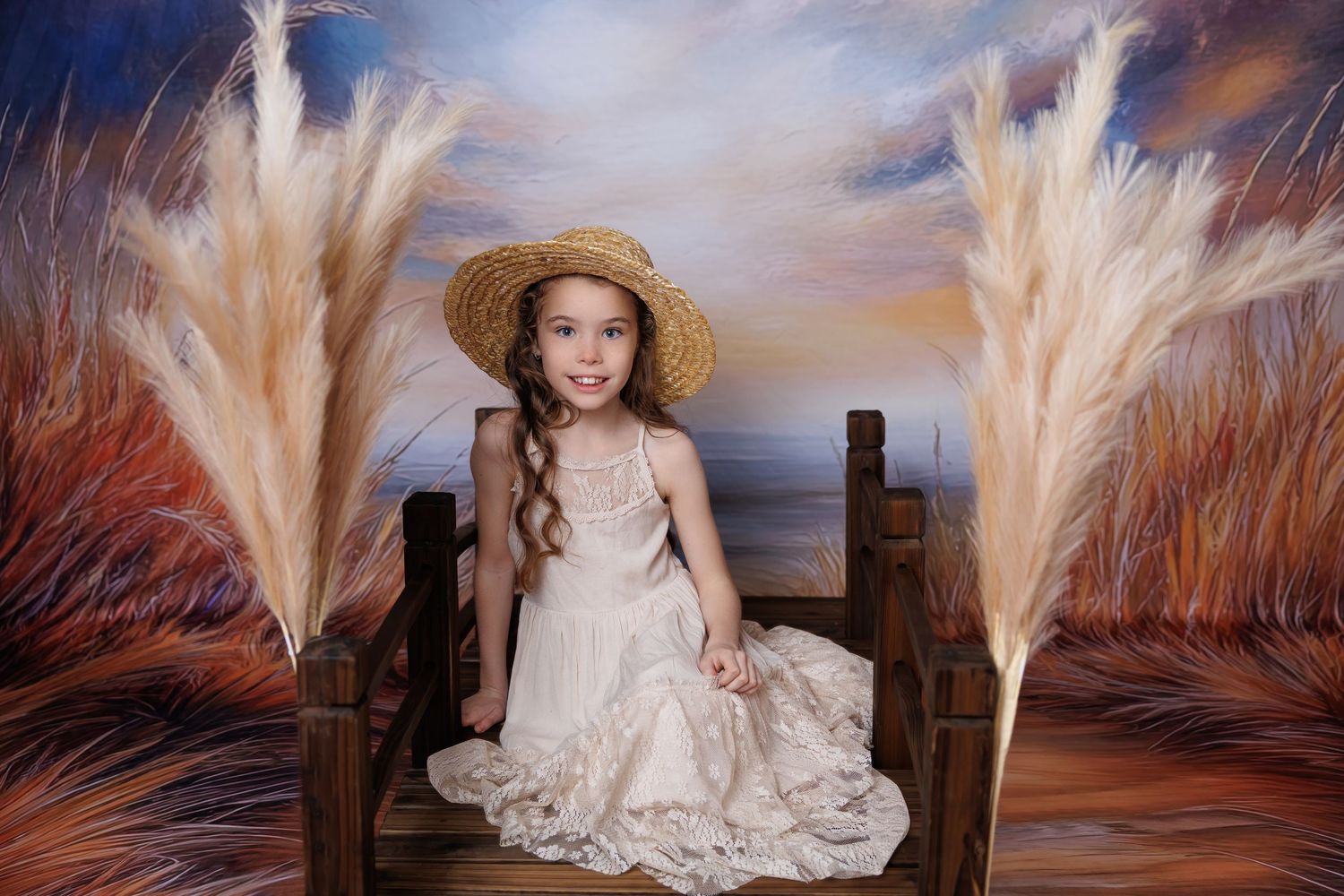 A young girl in a straw hat and cream dress sits on a wooden bench surrounded by pampas grass decor.