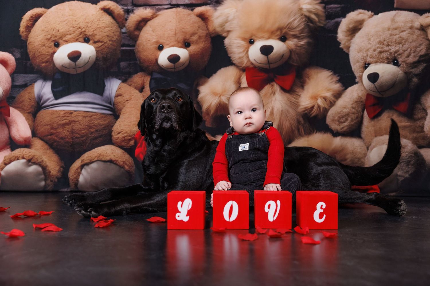 Baby in red outfit sits beside a black dog surrounded by teddy bears with LOVE blocks.