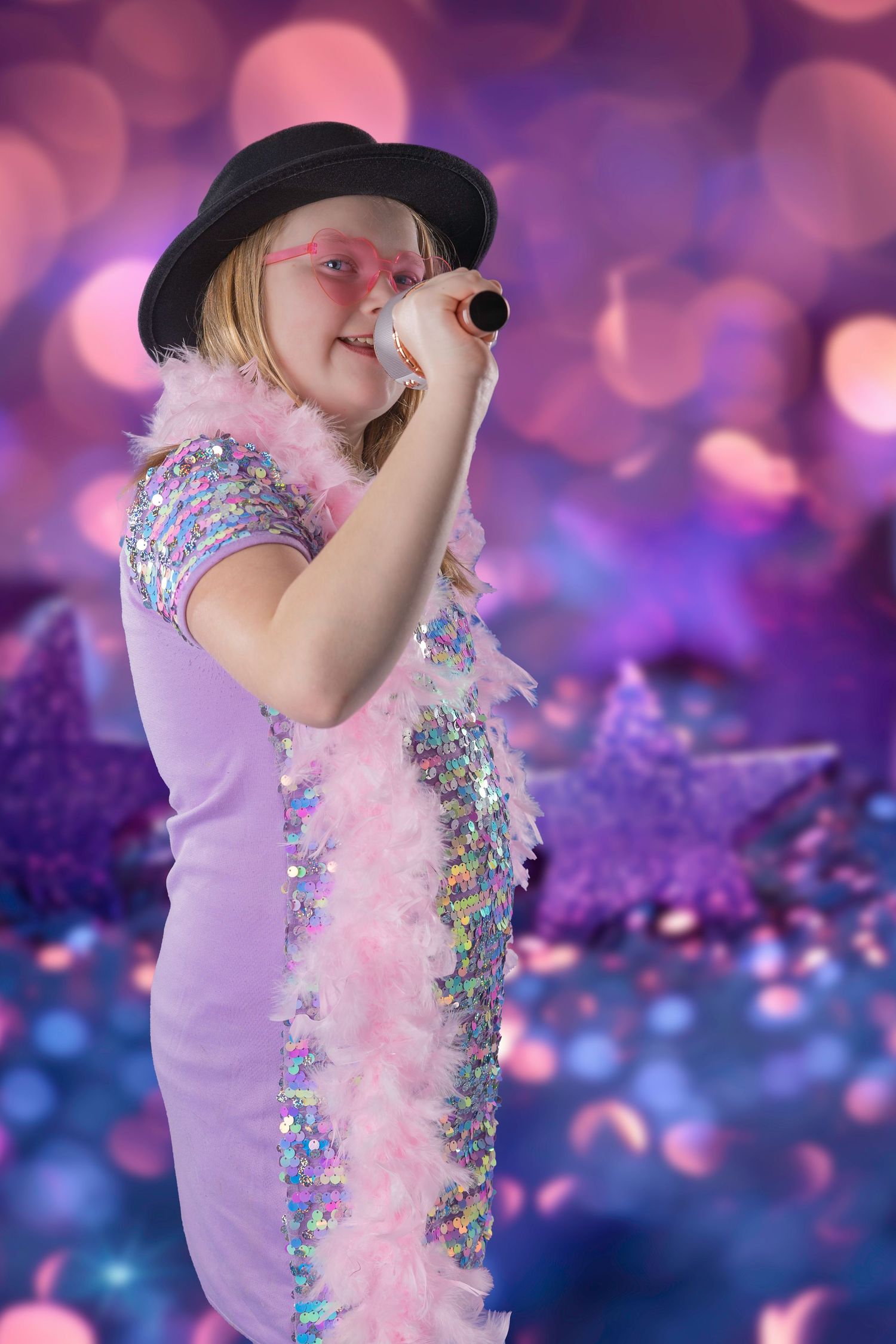 Young performer in pink heart sunglasses, black hat, and sequin dress sings into microphone on bokeh-lit stage.