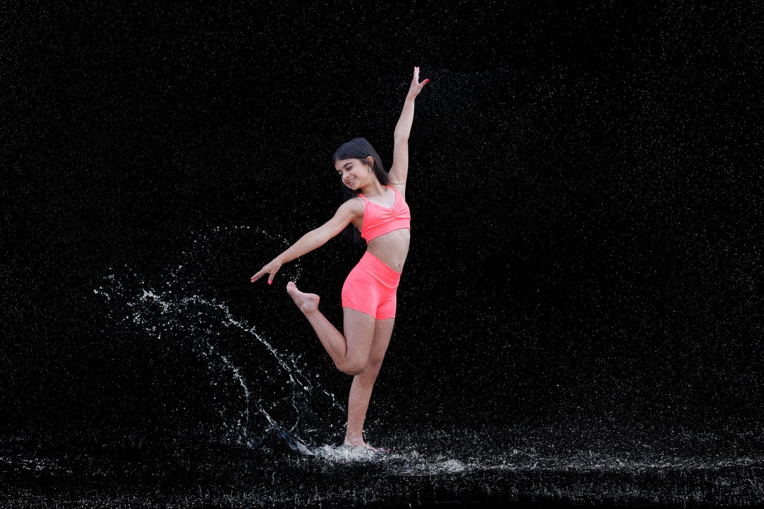 A young dancer in pink activewear kicks gracefully in rain splashes against a dark background.