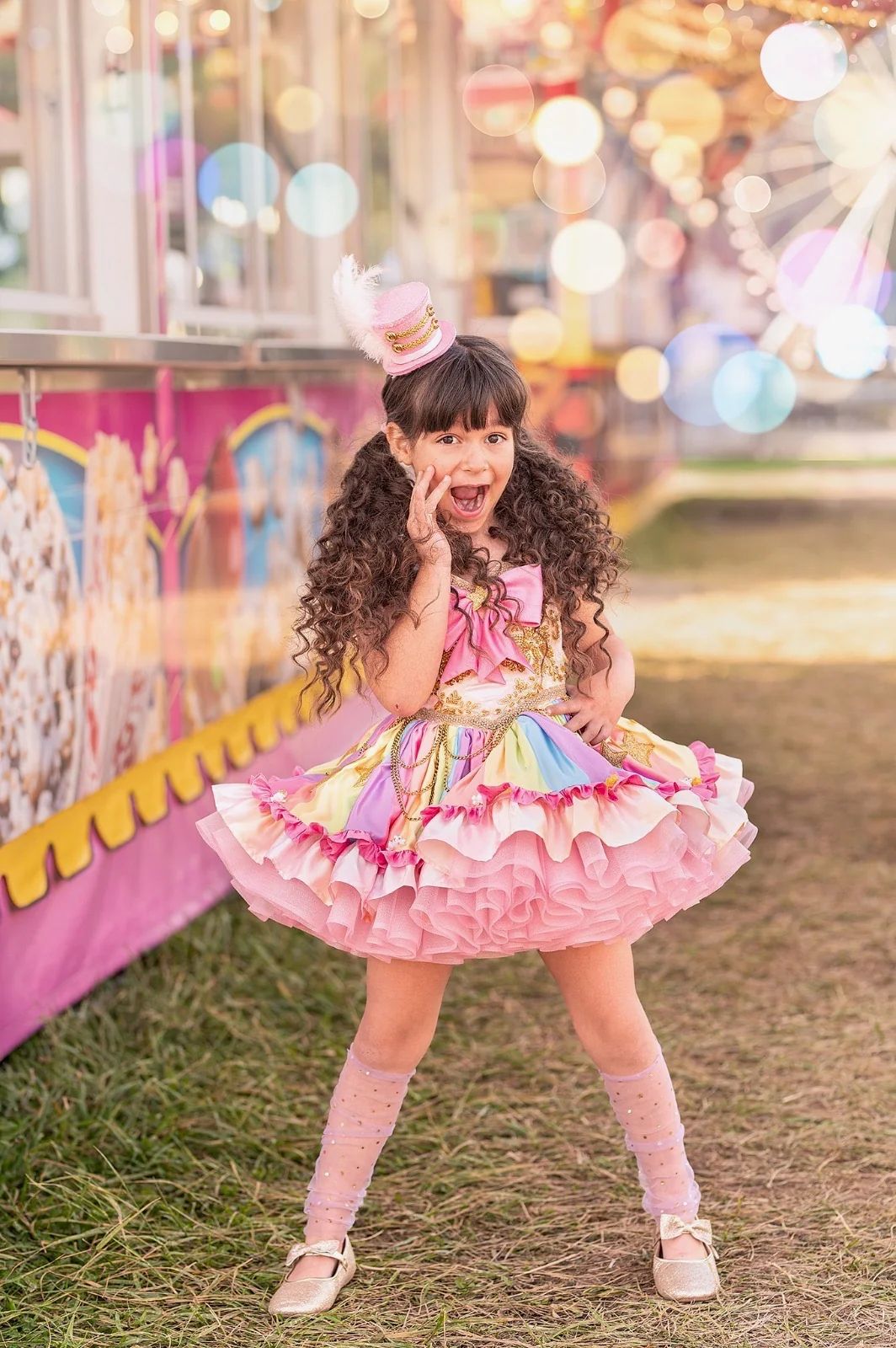 A young girl in a pink ruffled circus costume and mini top hat poses at a colorful carnival fair.