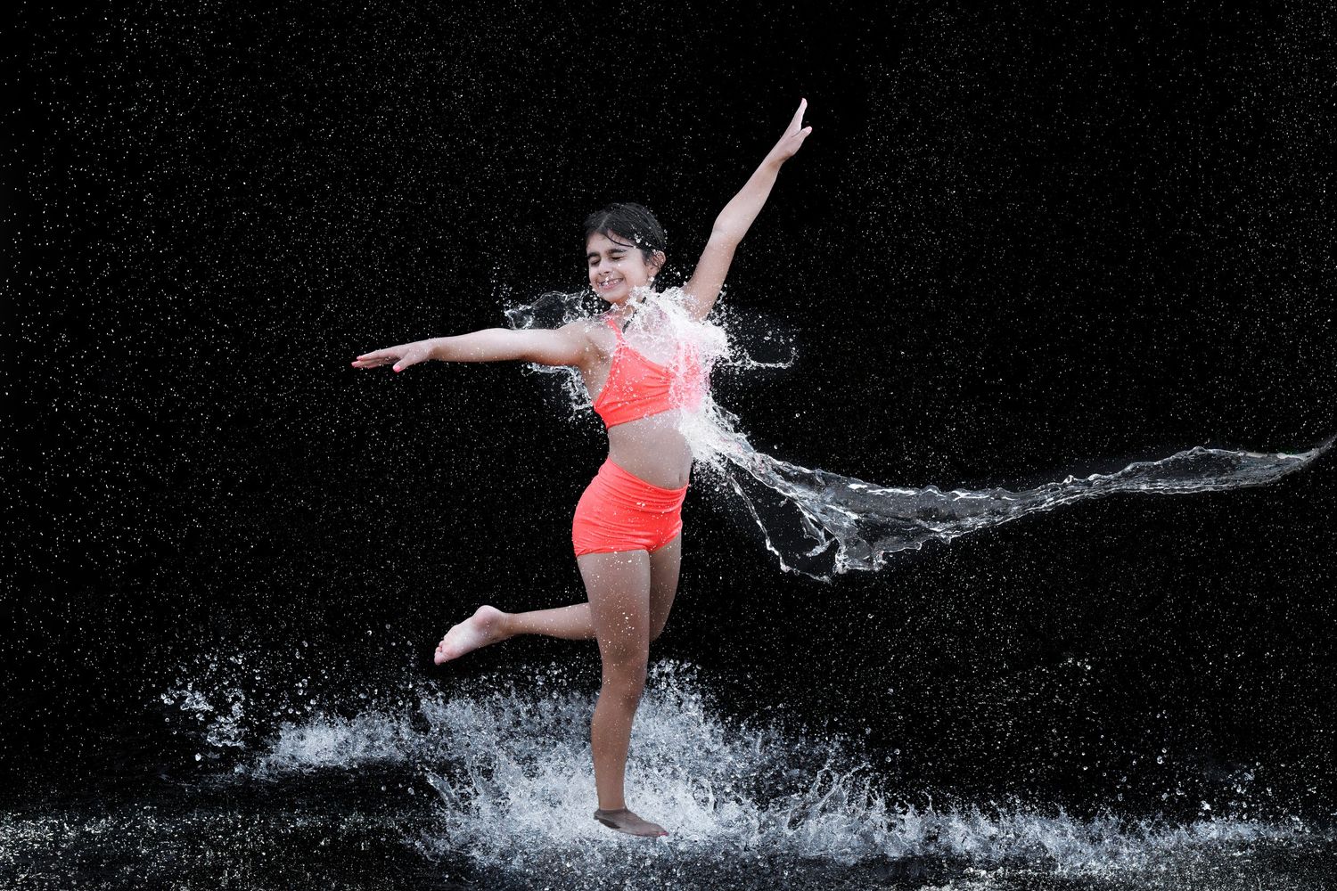 Young woman in orange bikini dancing joyfully in splashing water against a dark background.