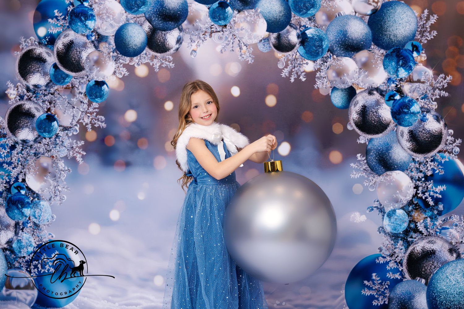 Girl in blue dress holds giant ornament under festive Christmas ball arch backdrop.