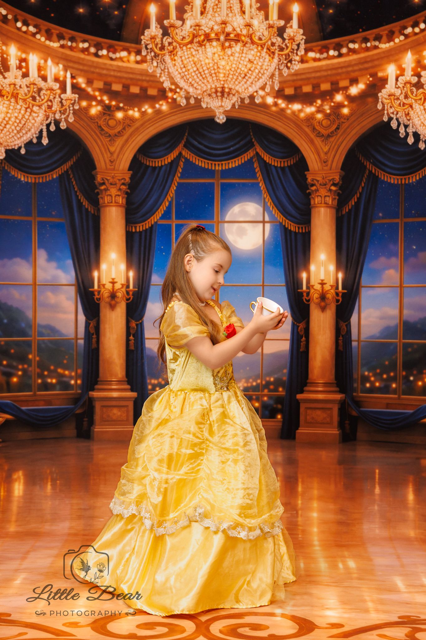 Young girl in yellow Belle gown holds teacup in enchanted ballroom with chandeliers and moonlit windows.
