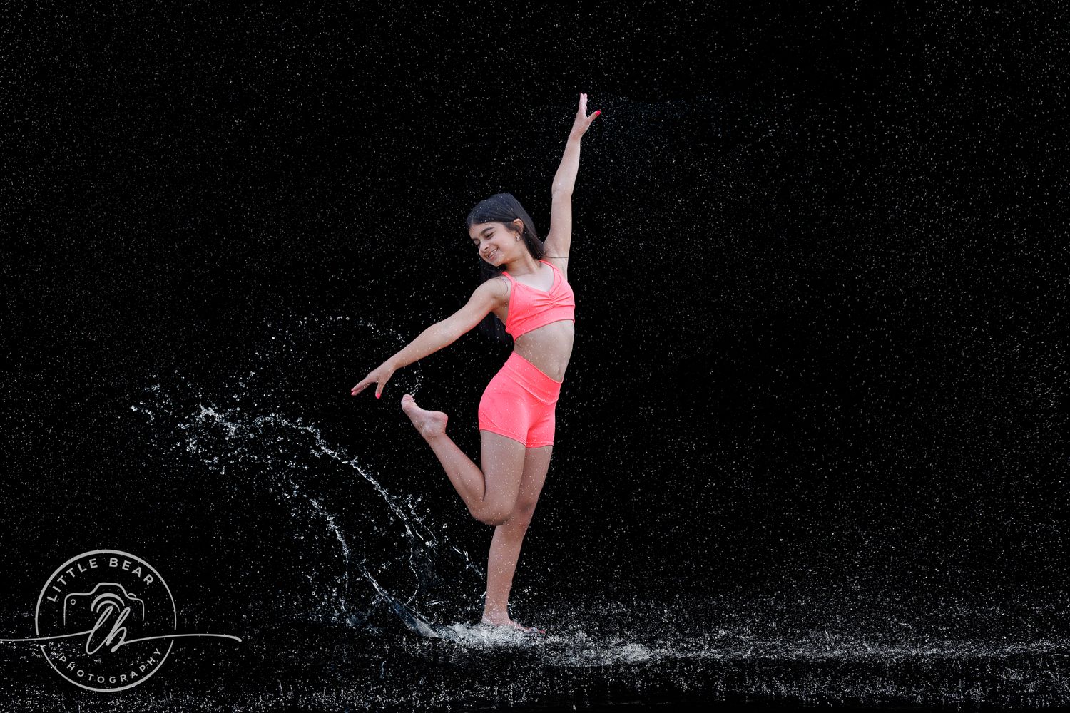 A young dancer in a pink outfit performs a graceful pose amid dynamic water splashes on a dark background.