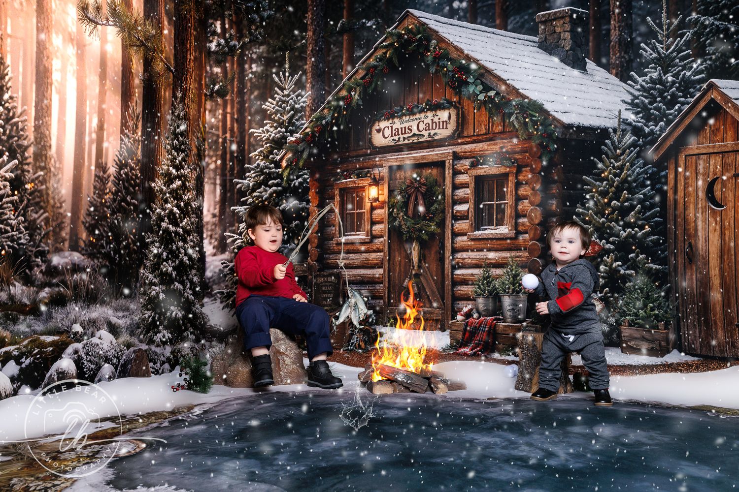 Two children sit by a campfire outside a snowy log cabin decorated for Christmas, surrounded by snow-covered pine trees.