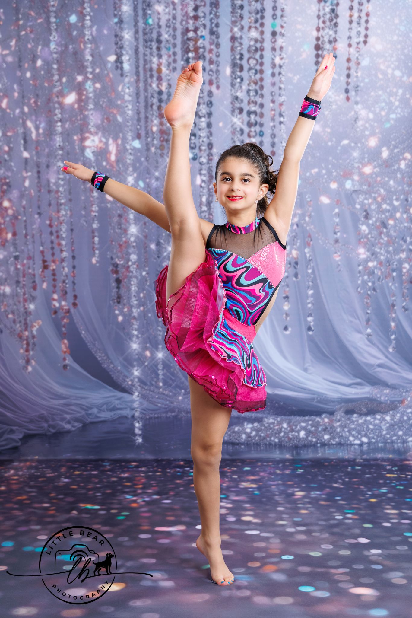 Young dancer in a vibrant pink and blue costume performs a high leg extension pose against a sparkling bokeh backdrop.