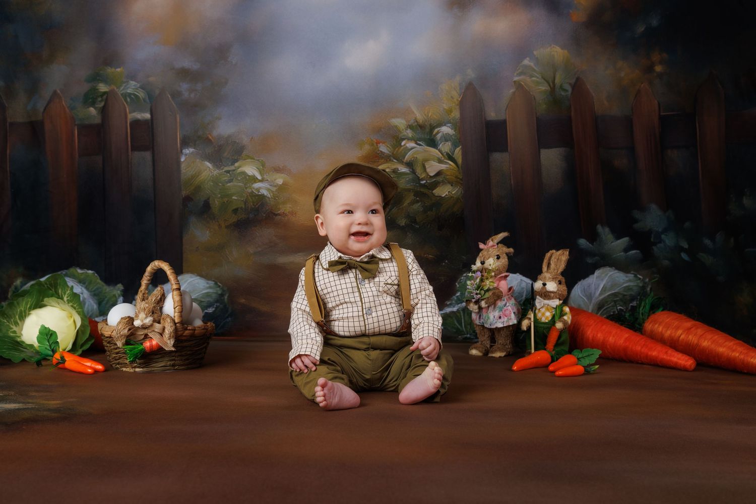 Happy smiling baby boy in suspenders sits among carrots and vegetables with Easter bunny themed backdrop.